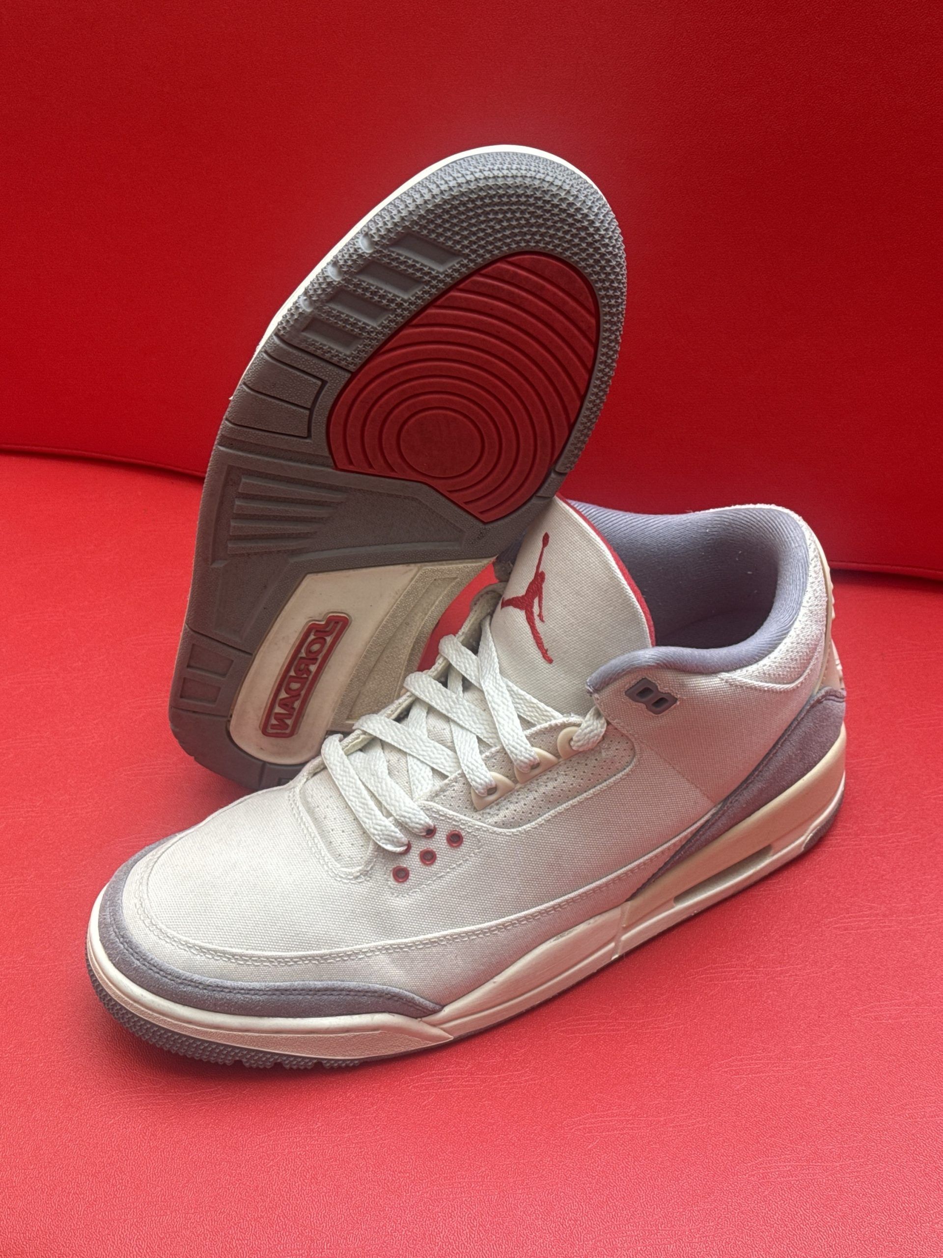Pair of light grey and red Air Jordan 3 sneakers on a red background, one shoe upside down.