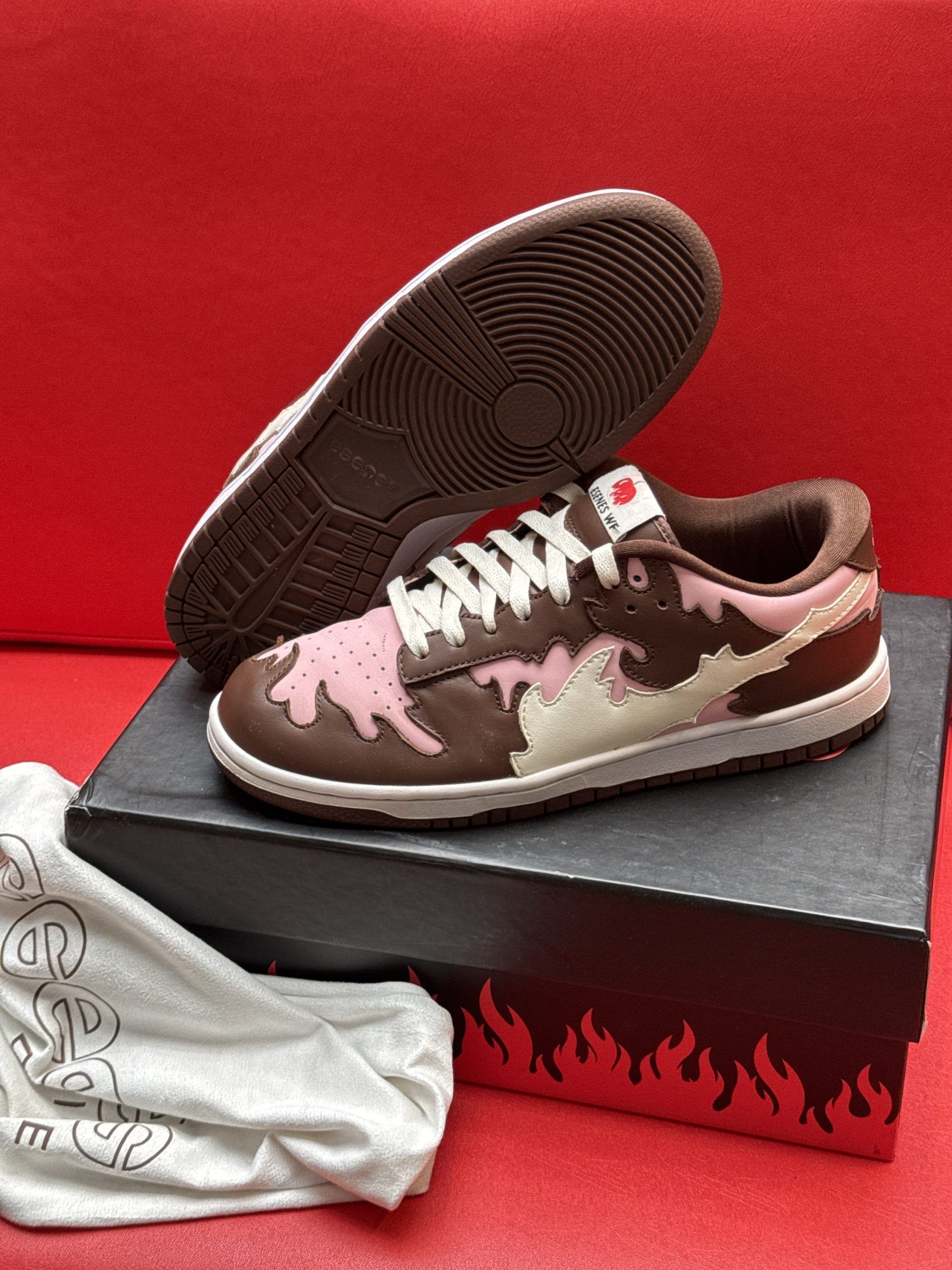 Brown and pink sneakers with flame design, sitting on a black box, and white bag.