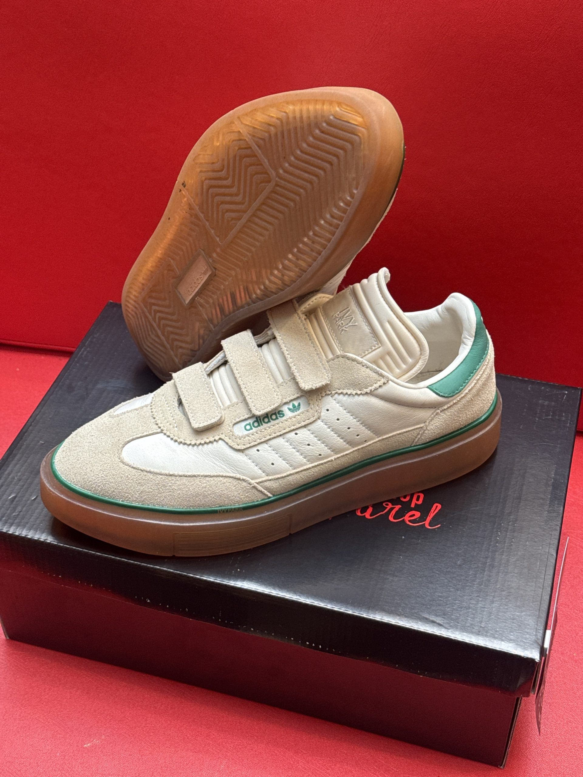 Beige Adidas sneaker with velcro straps, on a black box, with a red background.