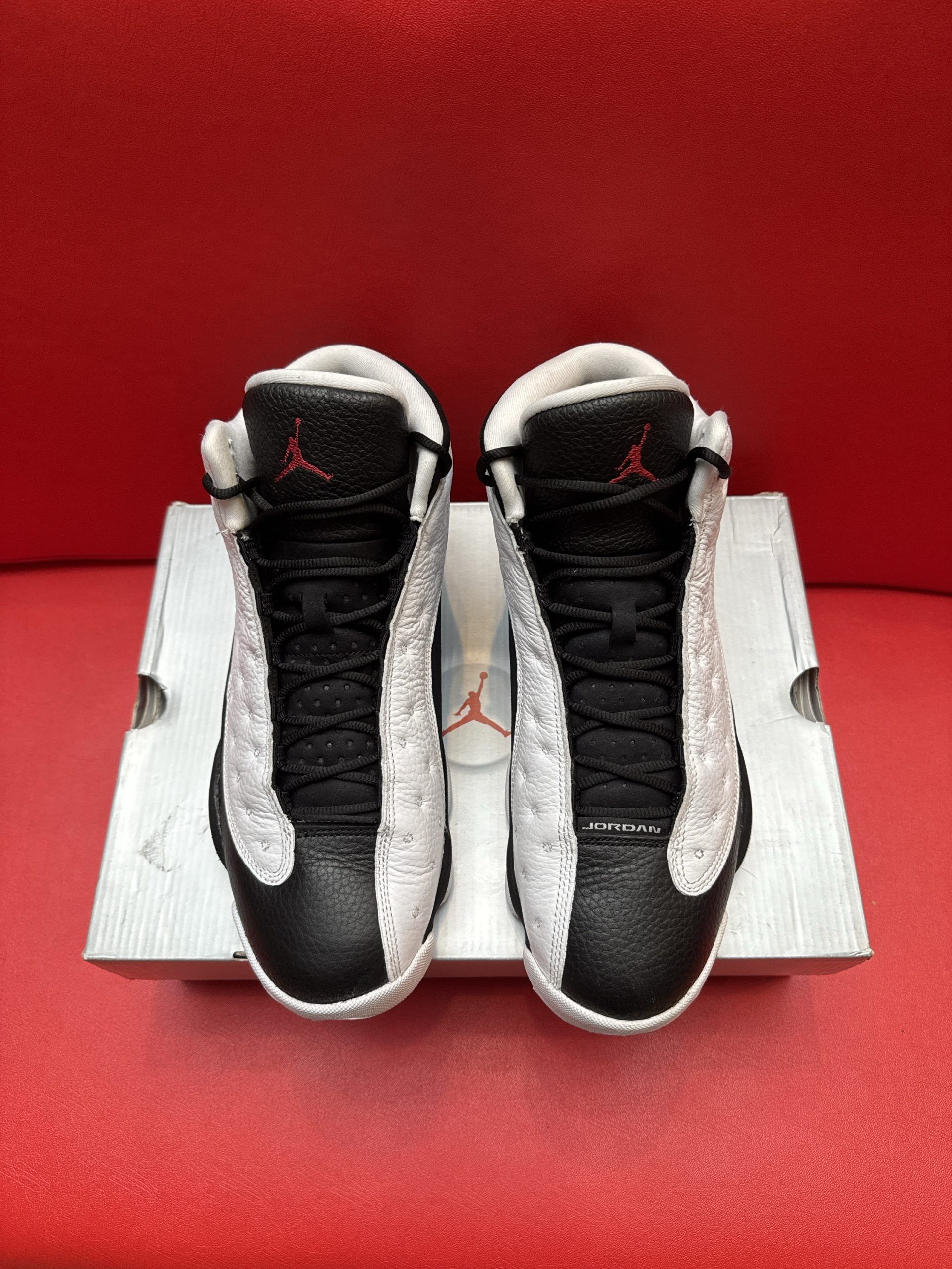 Air Jordan 13 sneakers, white and black with a red sole, resting on a shoebox against a red backdrop.