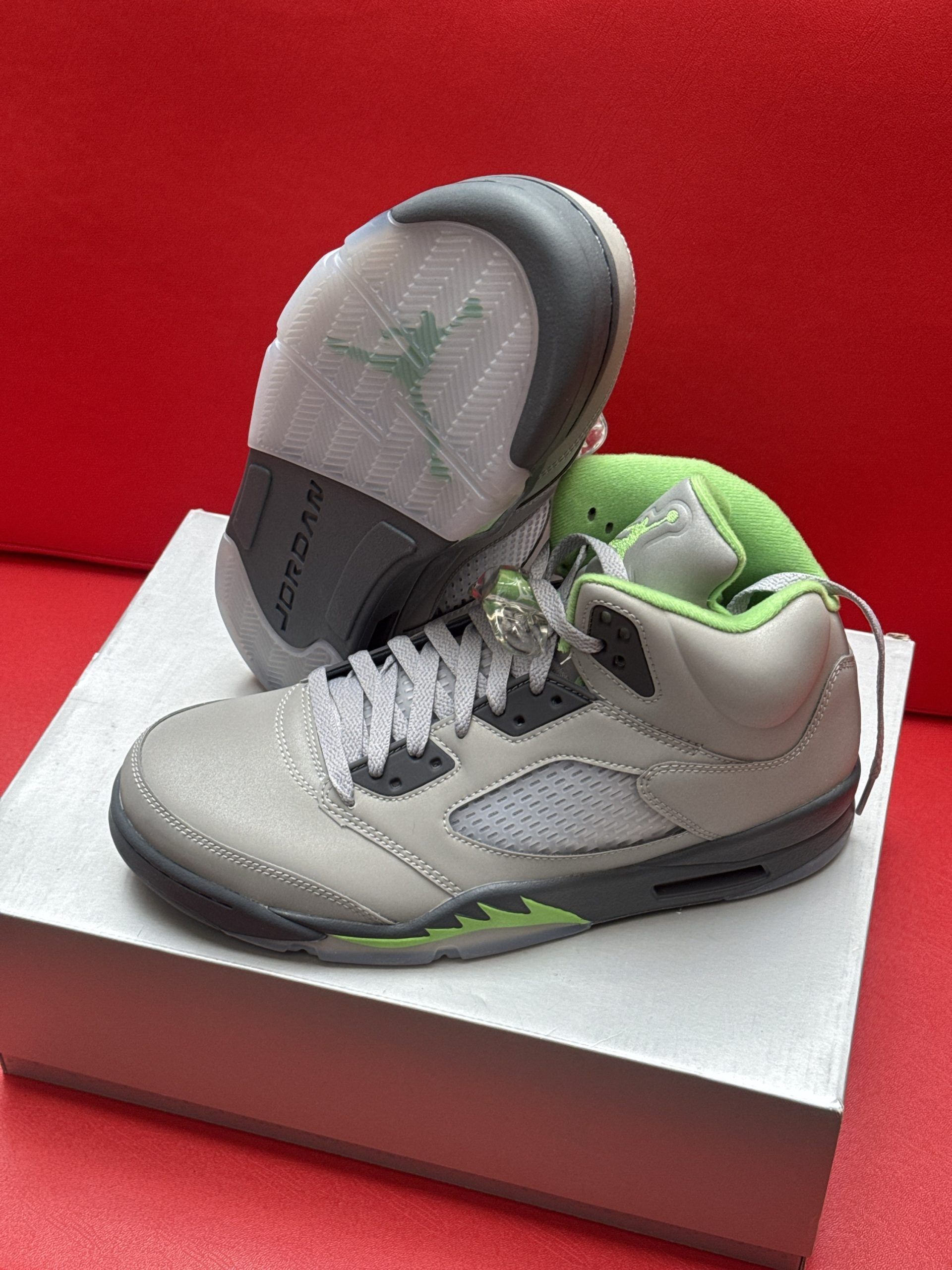 Gray, green, and black Air Jordan 5 sneakers on a white box against a red background.
