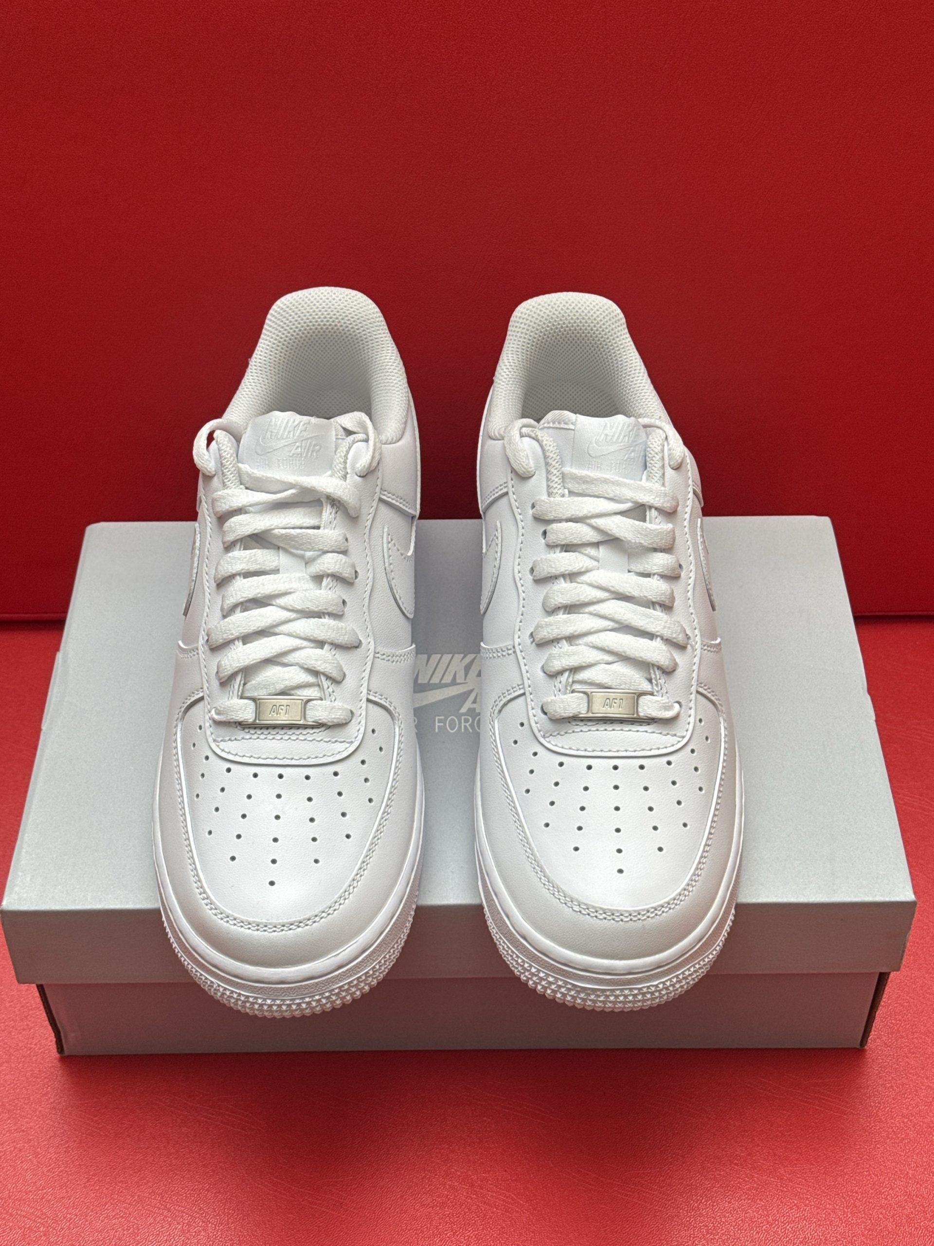 White Nike Air Force 1 sneakers on a box, sole up, against a red backdrop.