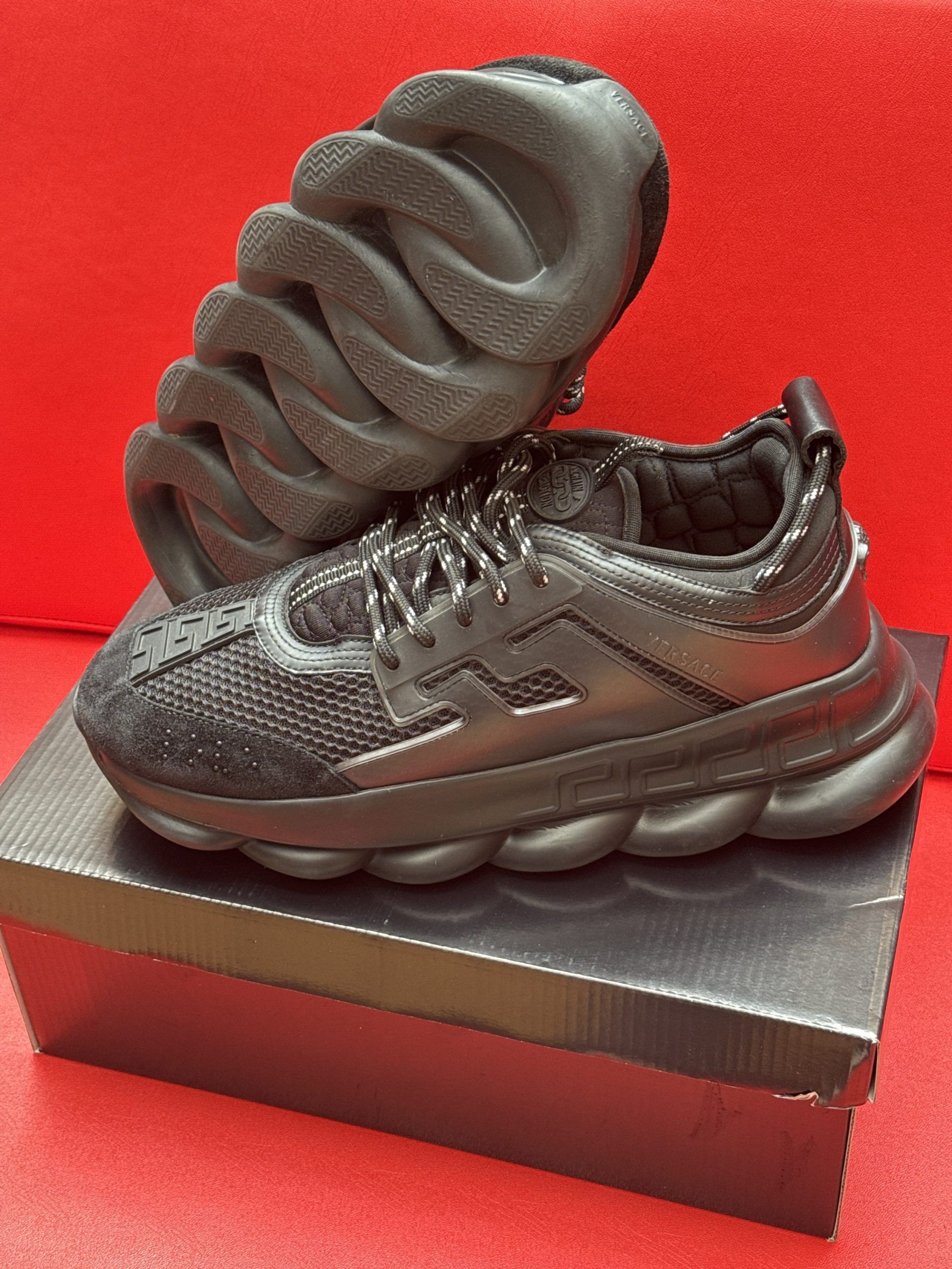 Black Versace Chain Reaction sneakers on a black box, with a red background.