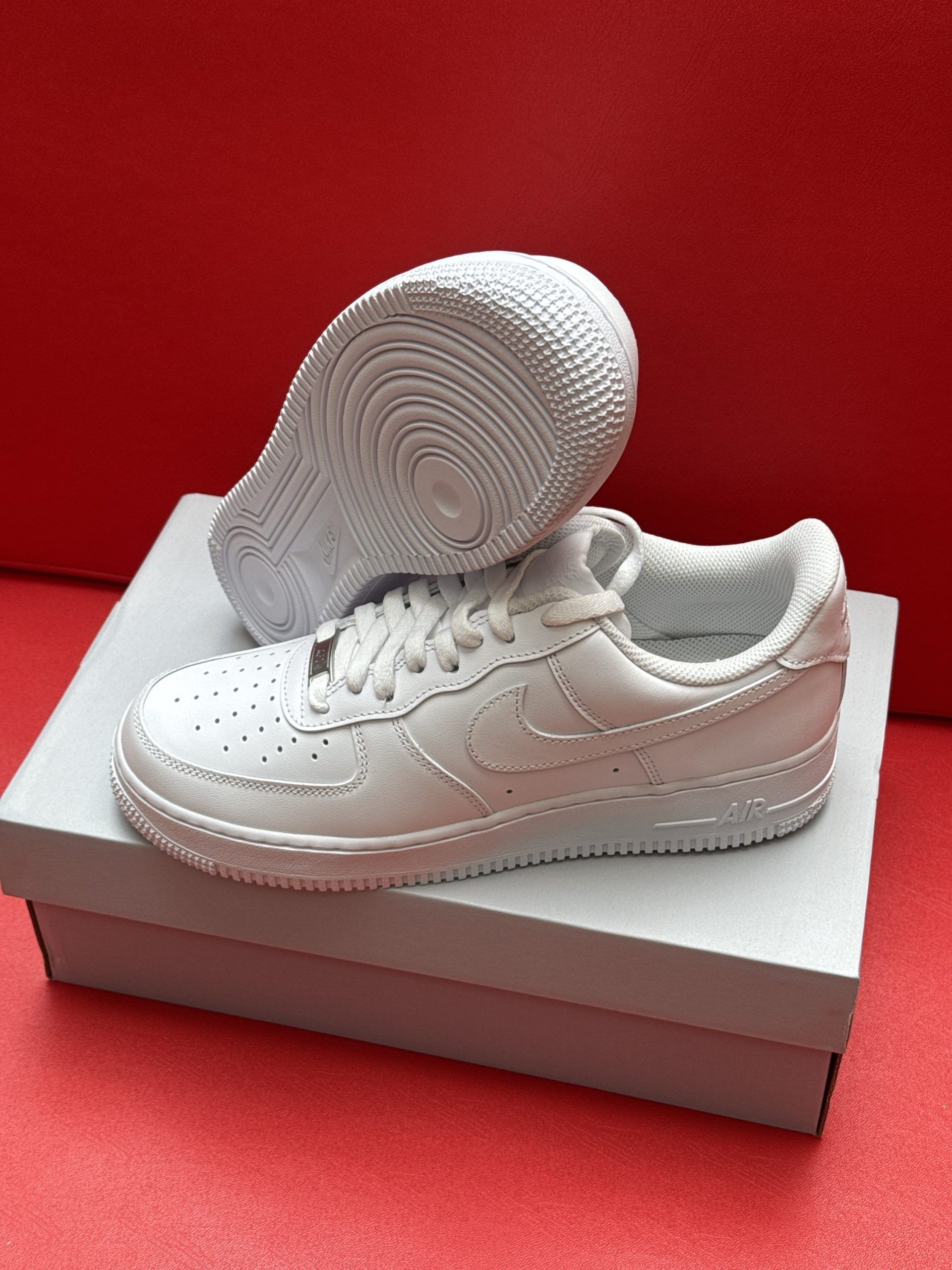 White Nike Air Force 1 sneakers on a box, sole up, against a red backdrop.