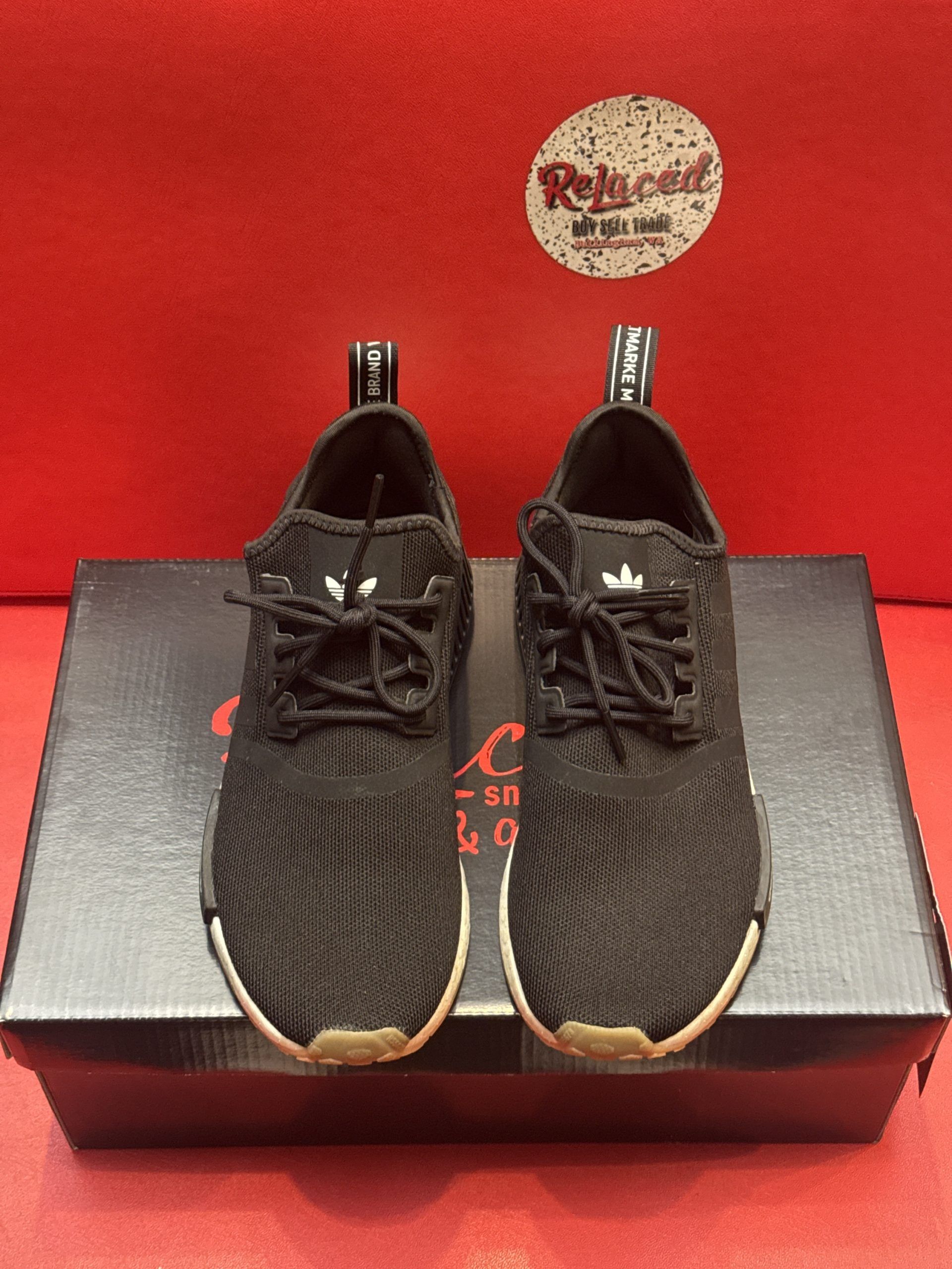 Black Adidas NMD sneakers with white and tan soles, on a black shoebox against a red background.