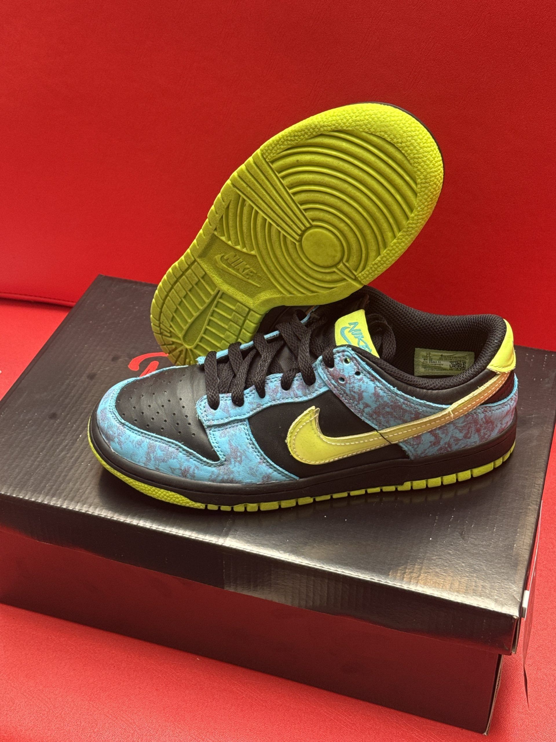 Nike Dunk low sneaker in black, blue, and yellow on a black shoe box with a red background.
