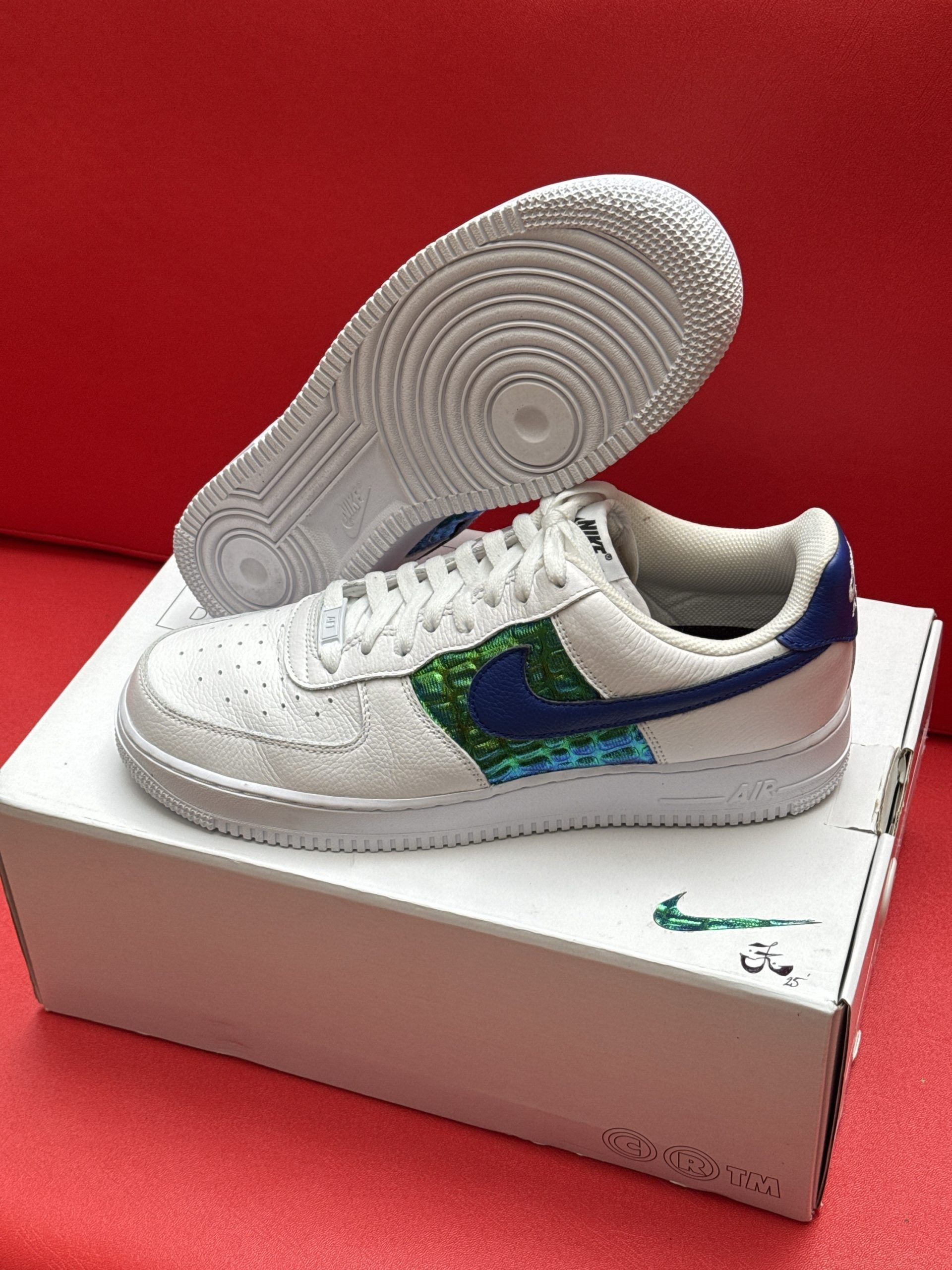 White Nike Air Force 1 sneakers with blue and green accents sit atop a white box against a red background.