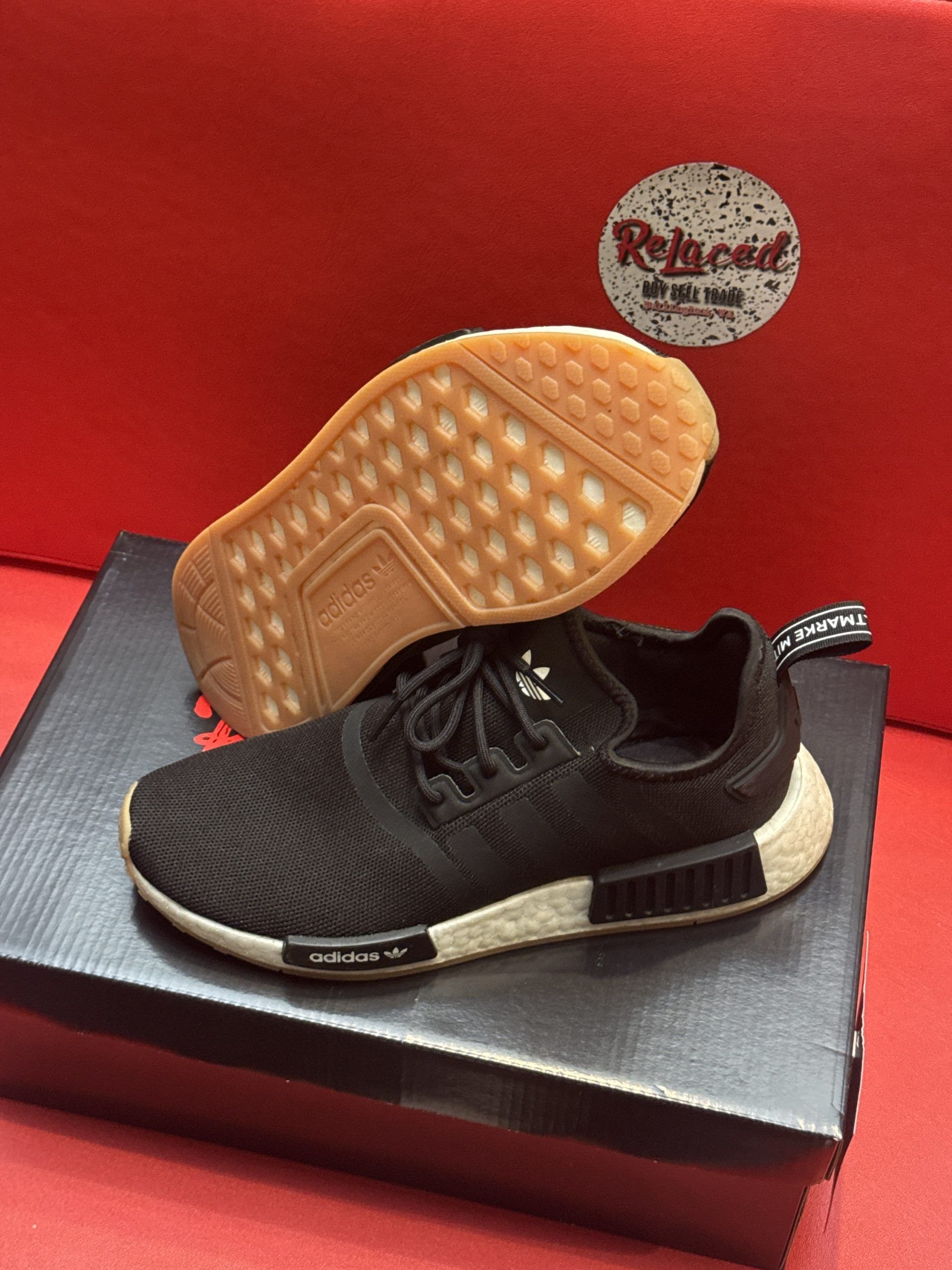 Black Adidas NMD sneakers with white and tan soles, on a black shoebox against a red background.