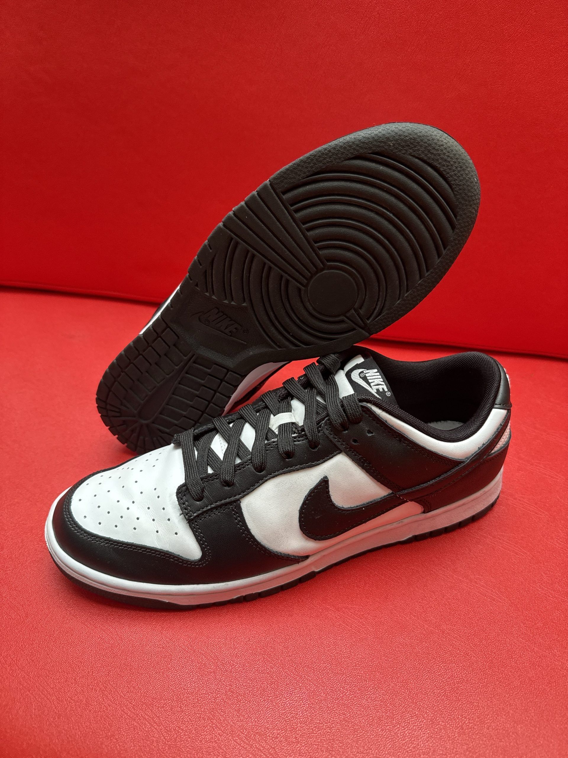 Black and white Nike Dunk Low sneakers, one upright, one showing the sole, on a red surface.