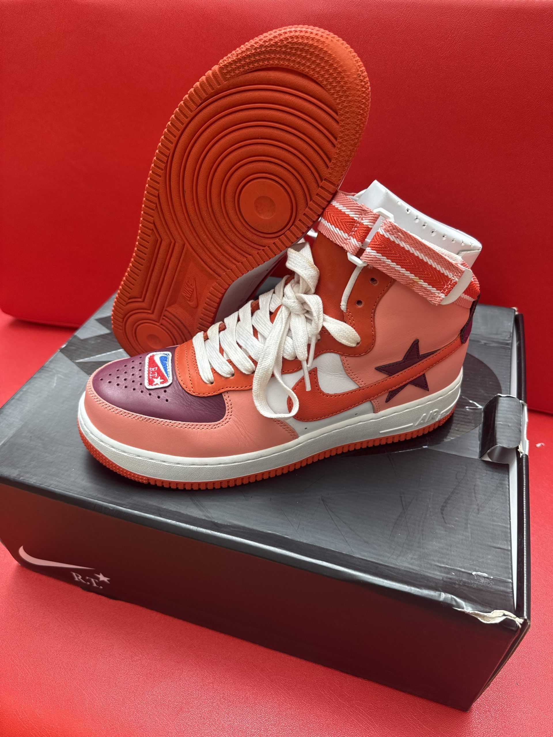 Red and pink Nike Air Force 1 high-top sneaker on top of a black Nike shoe box with a red sole.