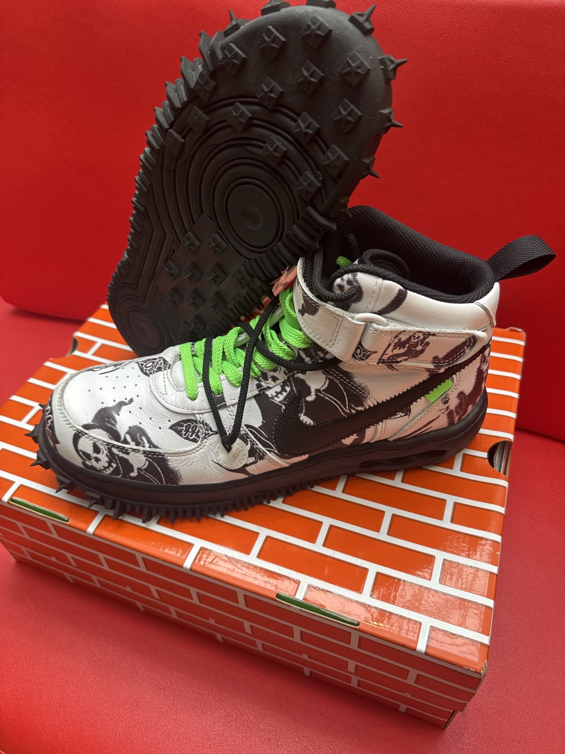 White and black patterned Nike sneaker with green laces, on a brick-patterned box, with black sole.