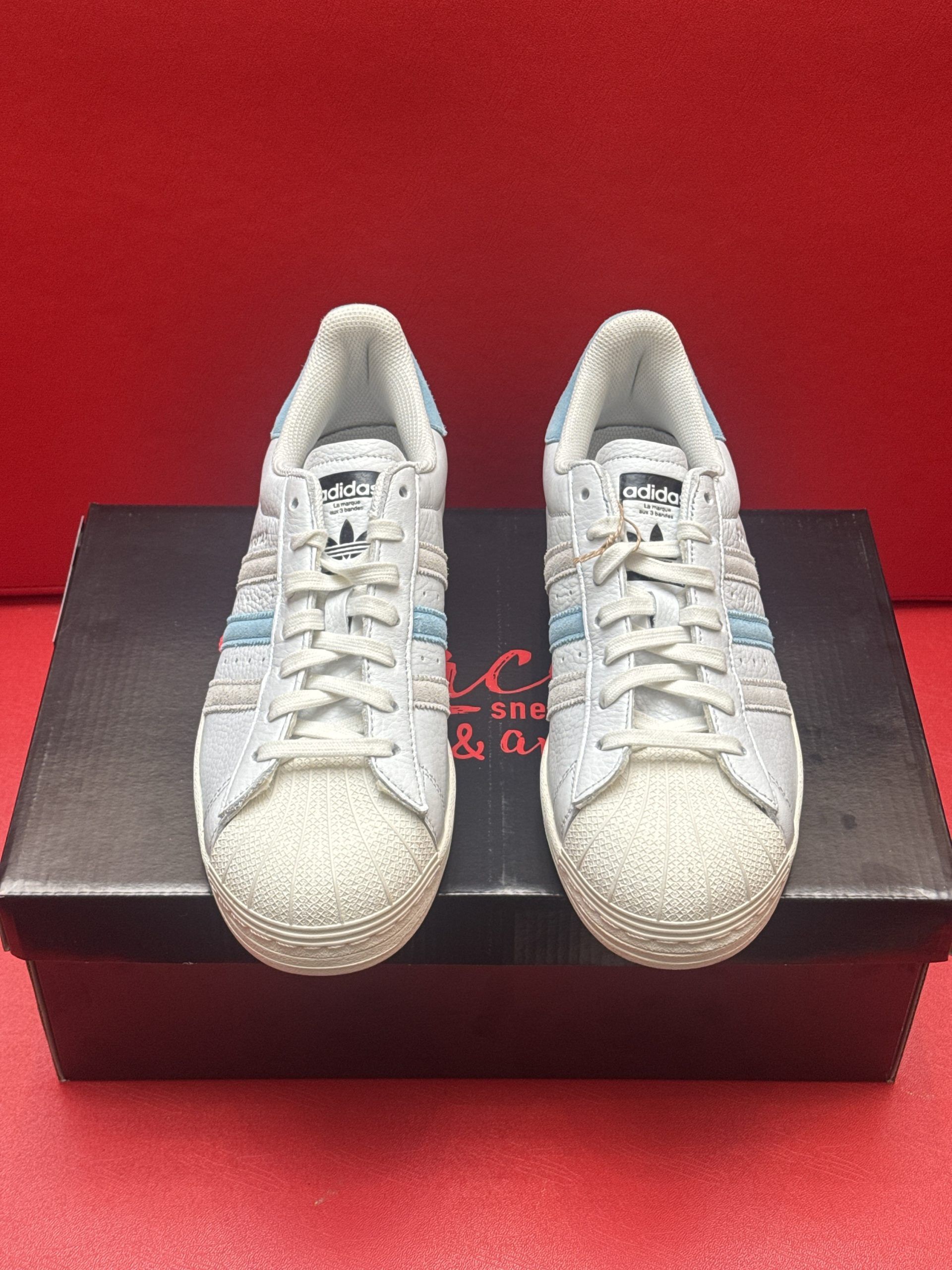 Light blue Adidas Superstar sneakers on a black box, one shoe lifted to show sole, set against a red background.