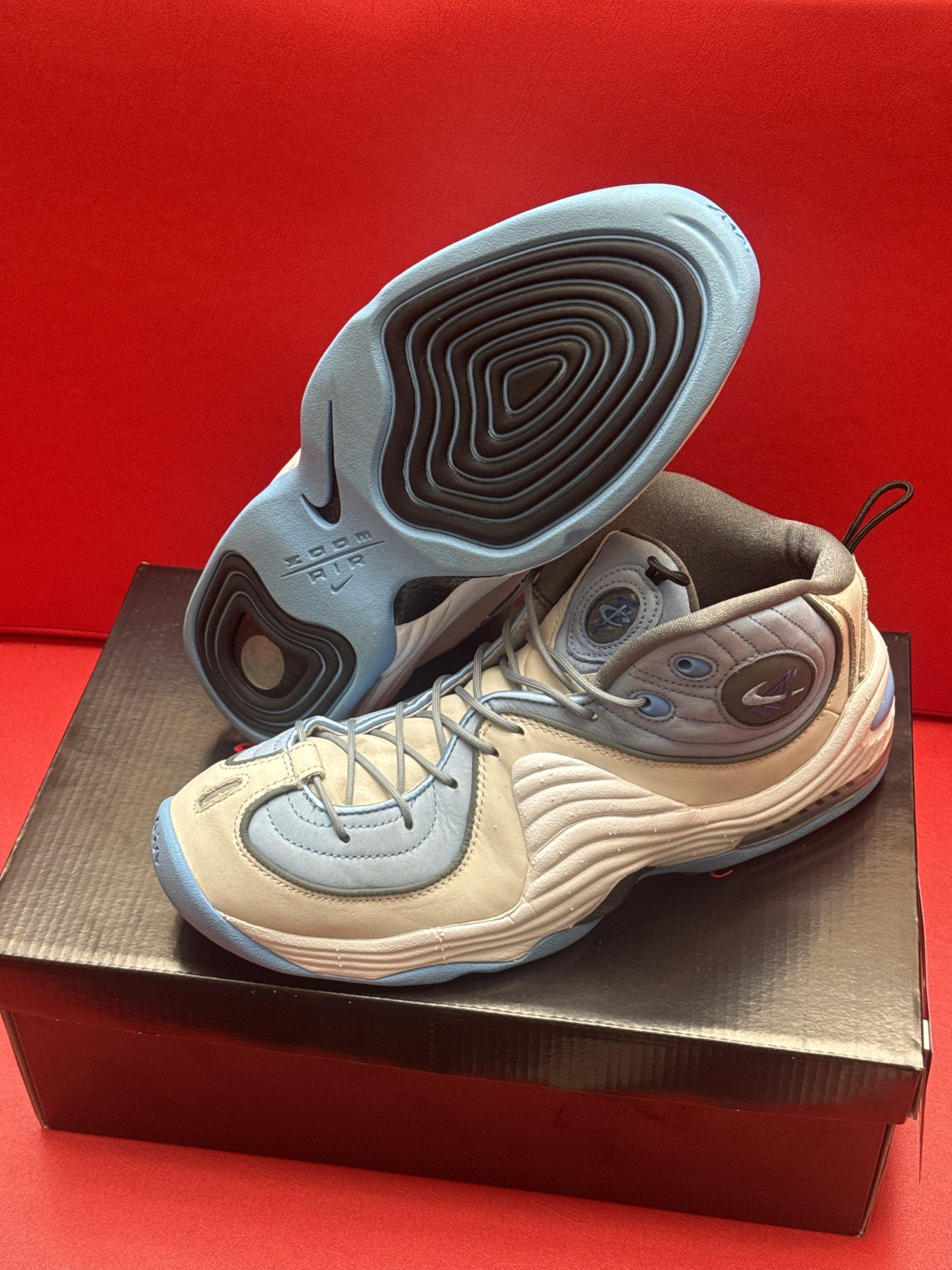 Light blue and gray Nike Penny Hardaway sneakers on a black box, red background.