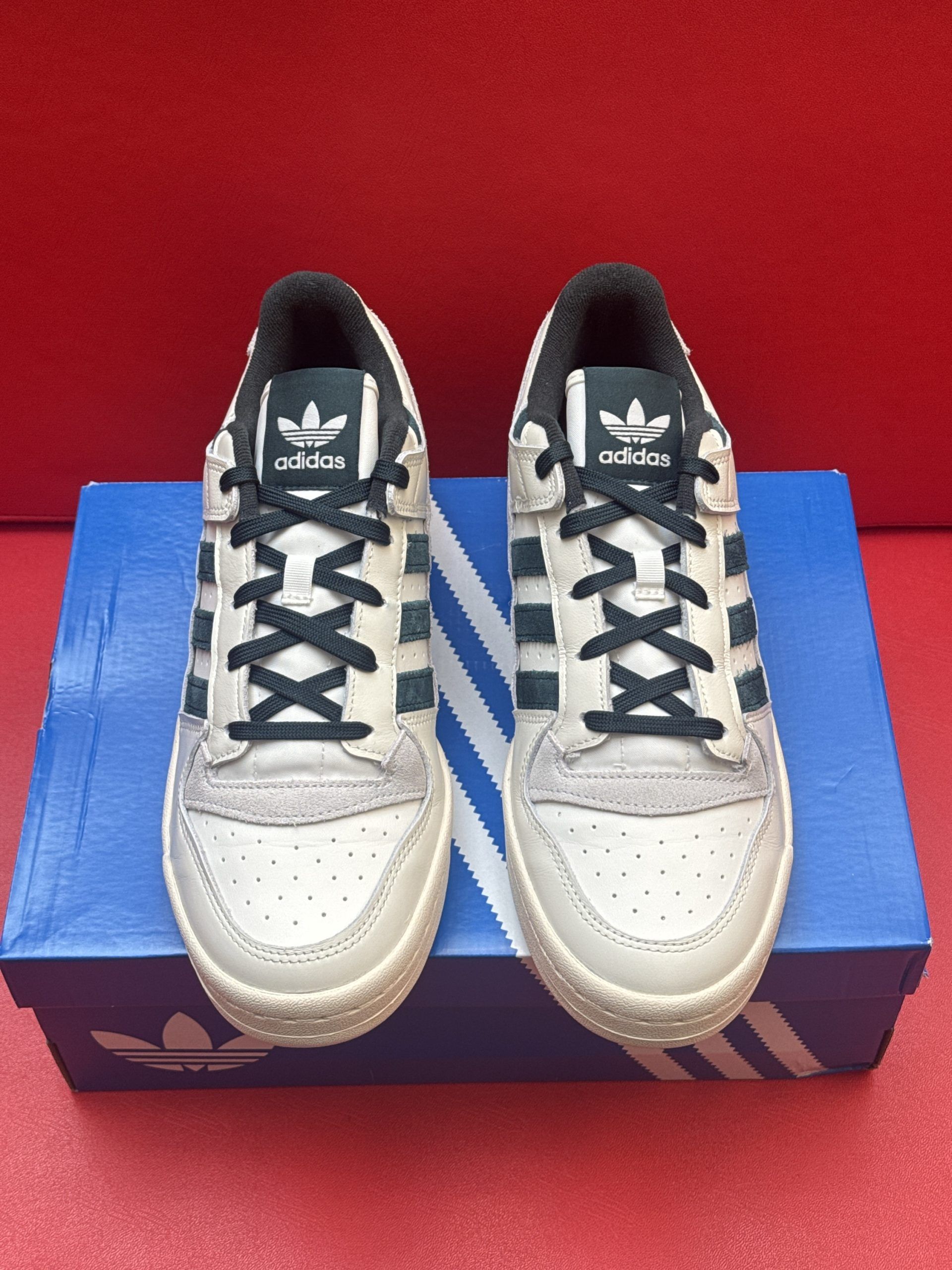Adidas Forum low sneakers in off-white with dark green stripes on the box against a red background.