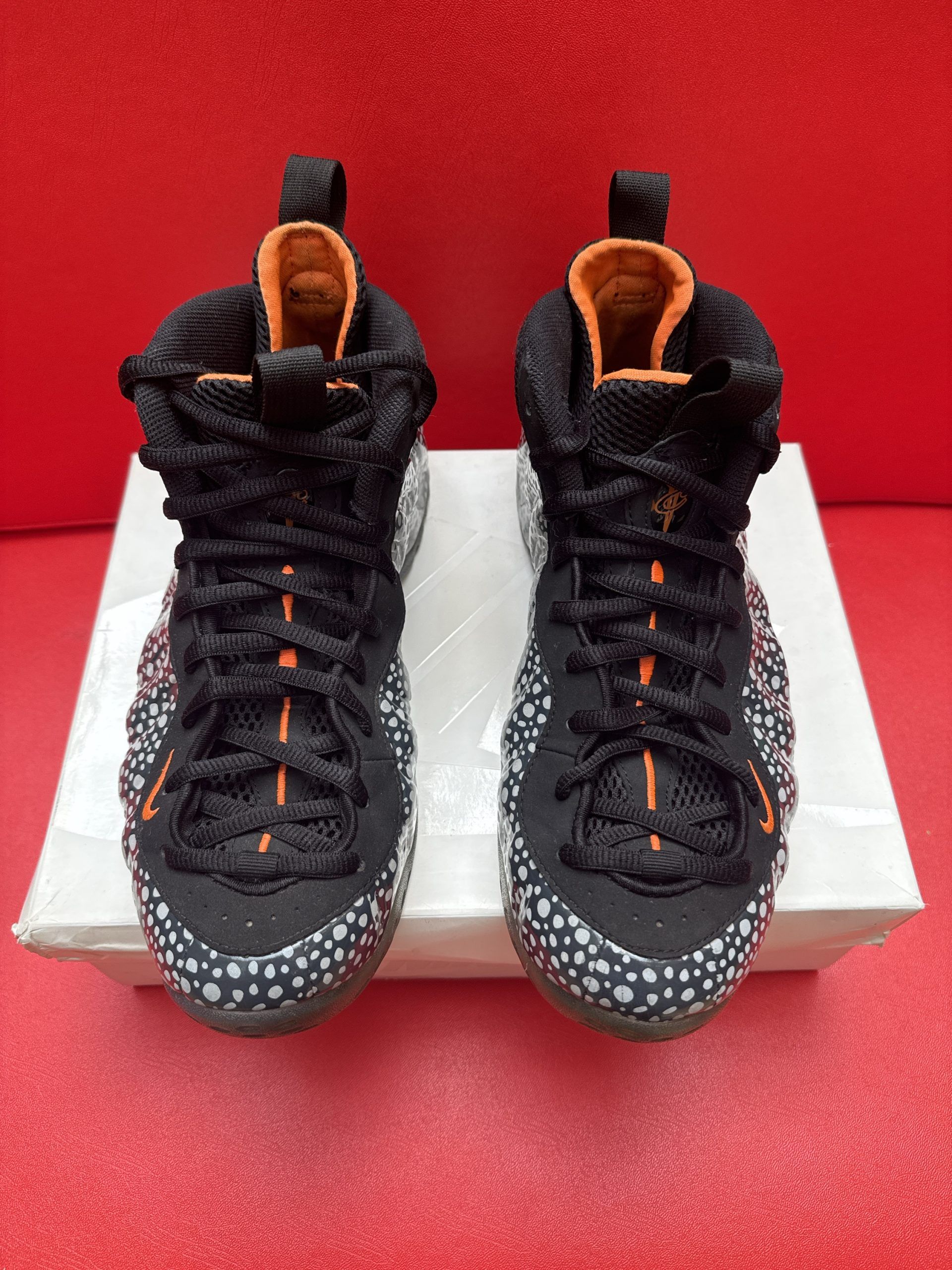 Black and speckled Nike Foamposite sneakers, on a white box, against a red backdrop.