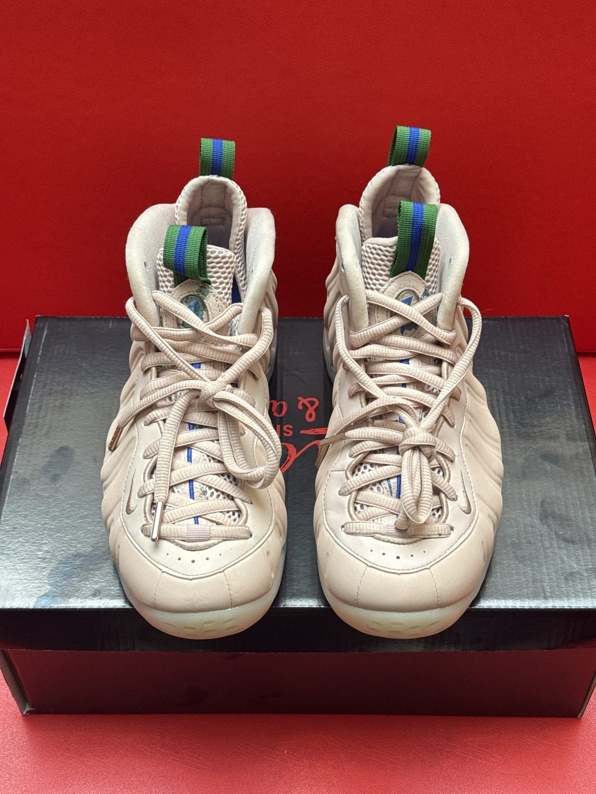 Tan Nike Foamposite sneakers on a black shoebox, sole up.  Green and blue details, red background.