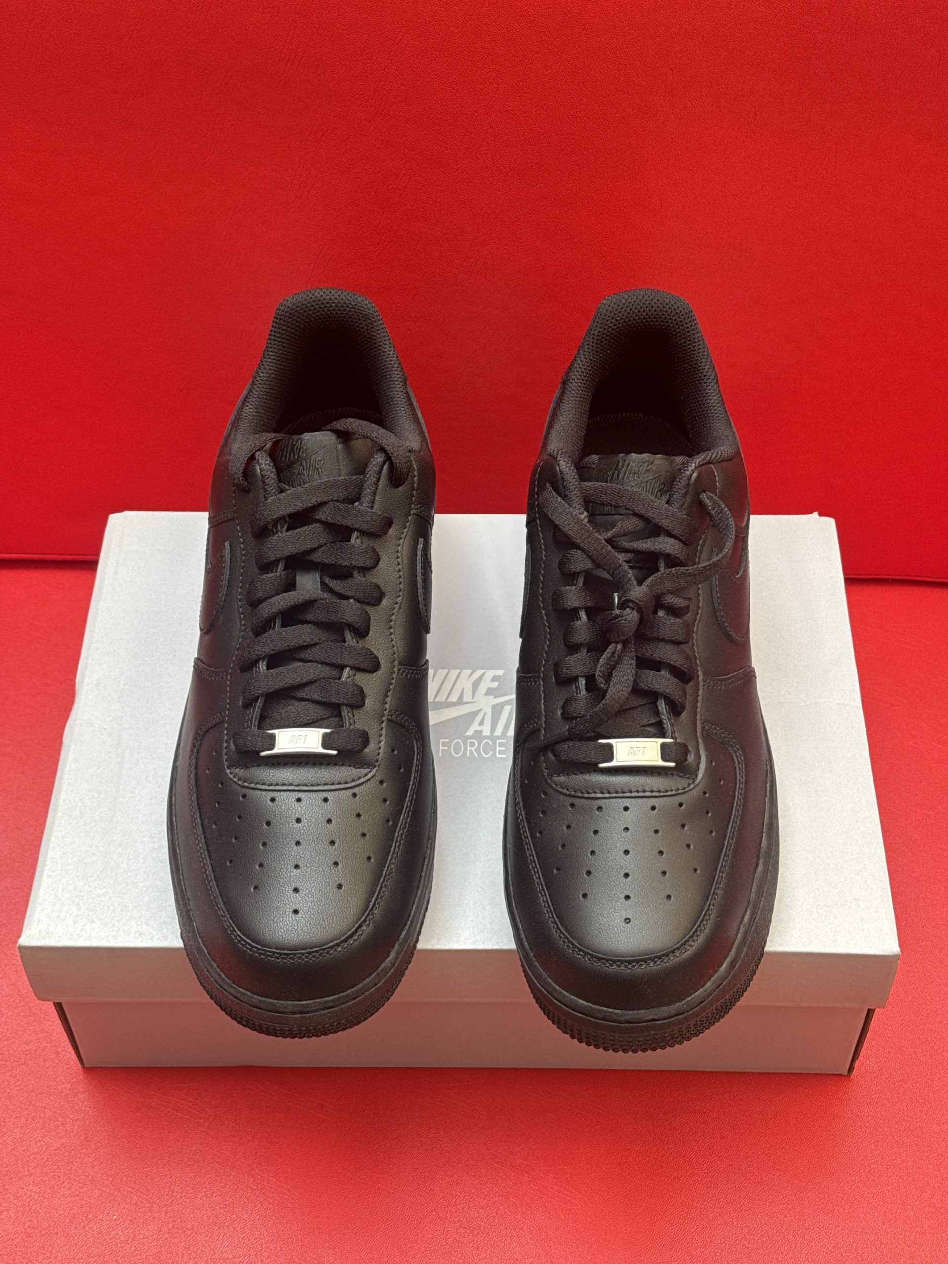 Black Nike Air Force 1 sneakers on a white box, set against a red background. The sole is visible.