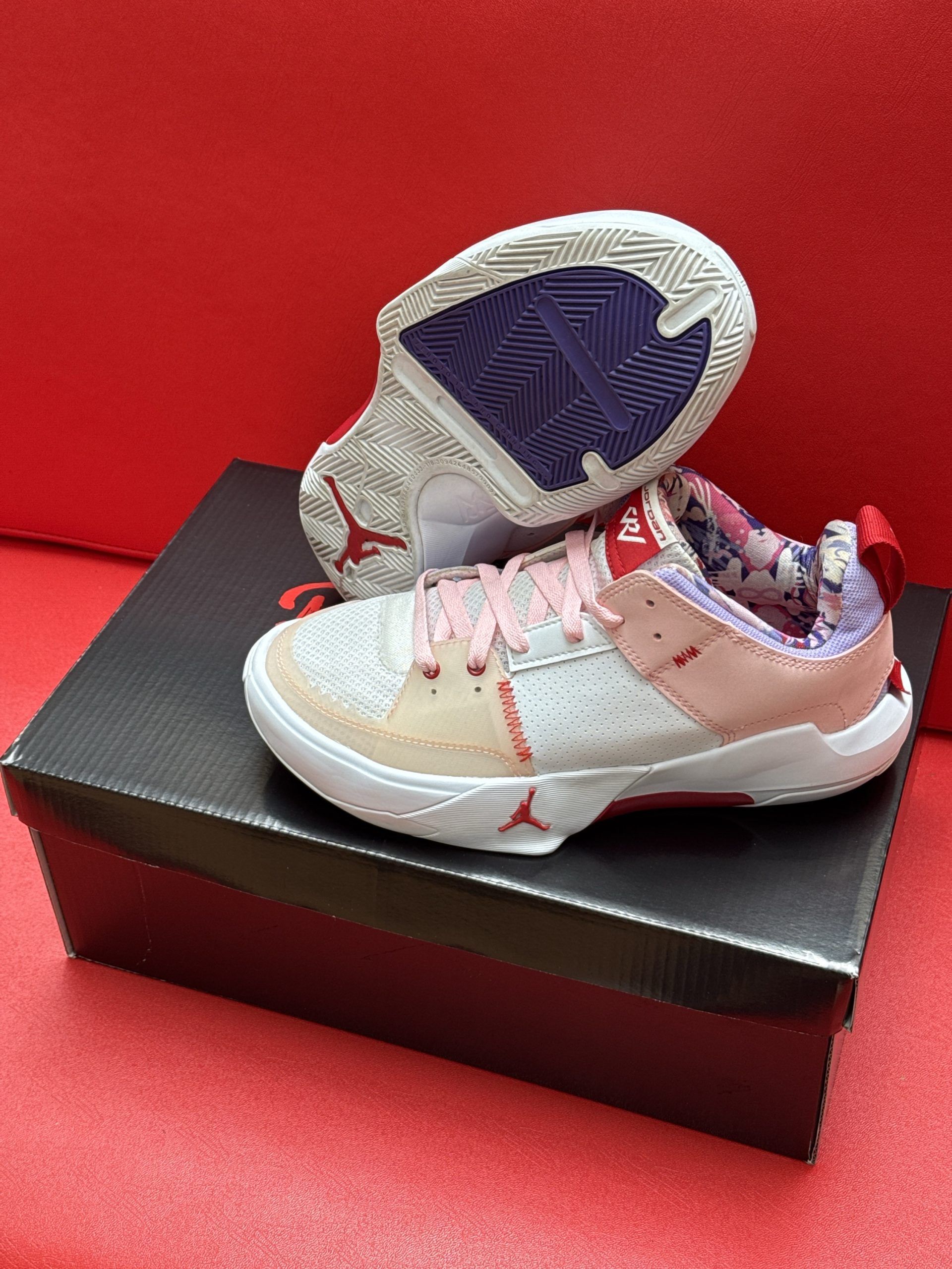 White, pink, and red Jordan sneakers on a black shoe box; red background.