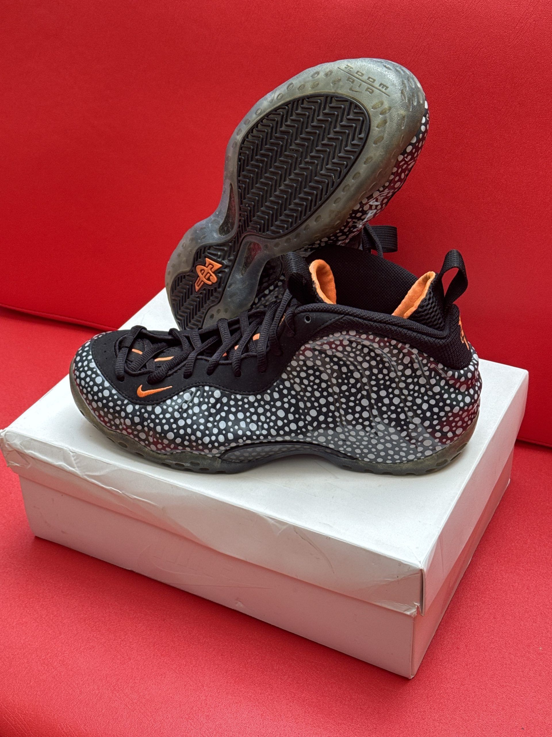 Black and speckled Nike Foamposite sneakers, on a white box, against a red backdrop.