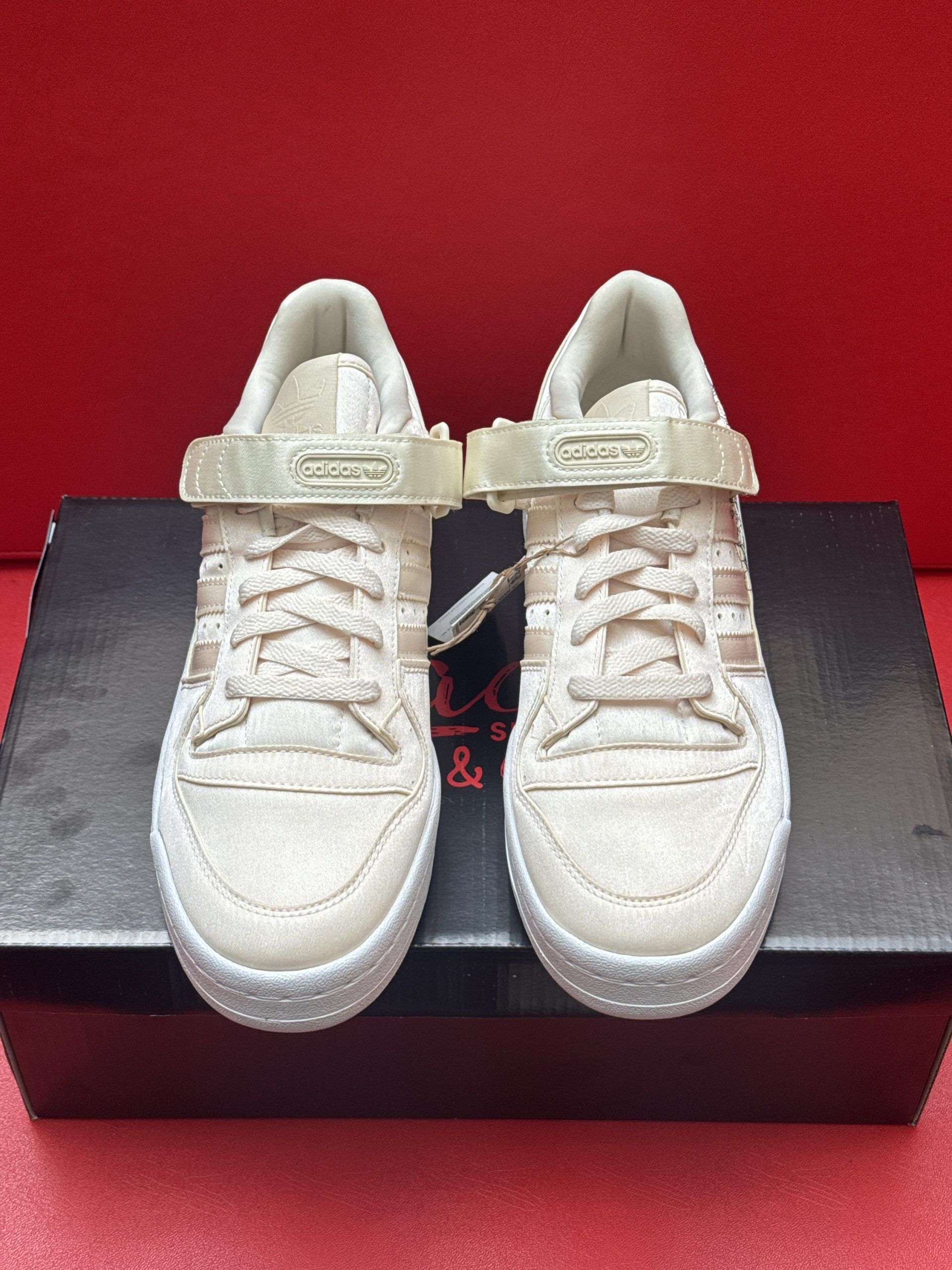 Pair of beige Adidas Forum Low sneakers on a black box, one shoe sole up, on a red surface.