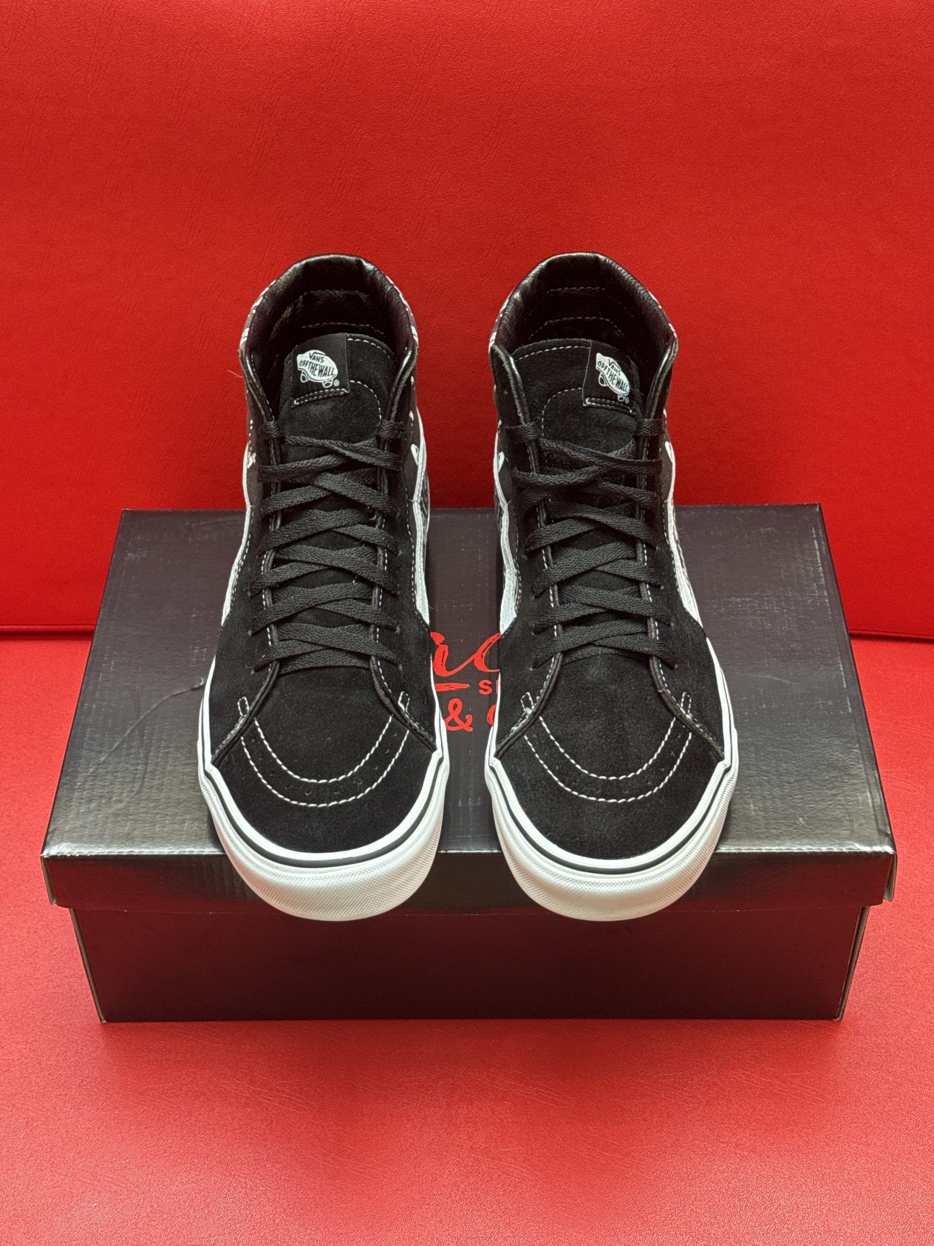 Black and white Vans SK8-Hi sneakers with a paisley pattern sit on a black shoebox against a red background.