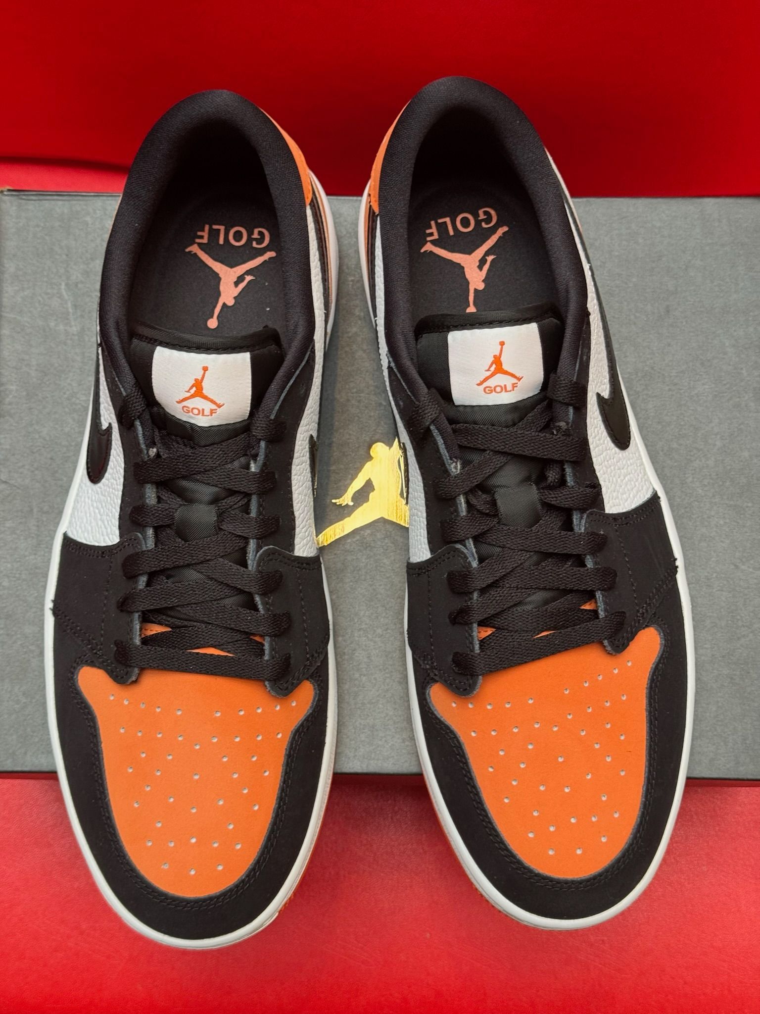 Orange, black, and white Air Jordan 1 Low sneaker displayed with the sole above the shoe on a box, red backdrop.