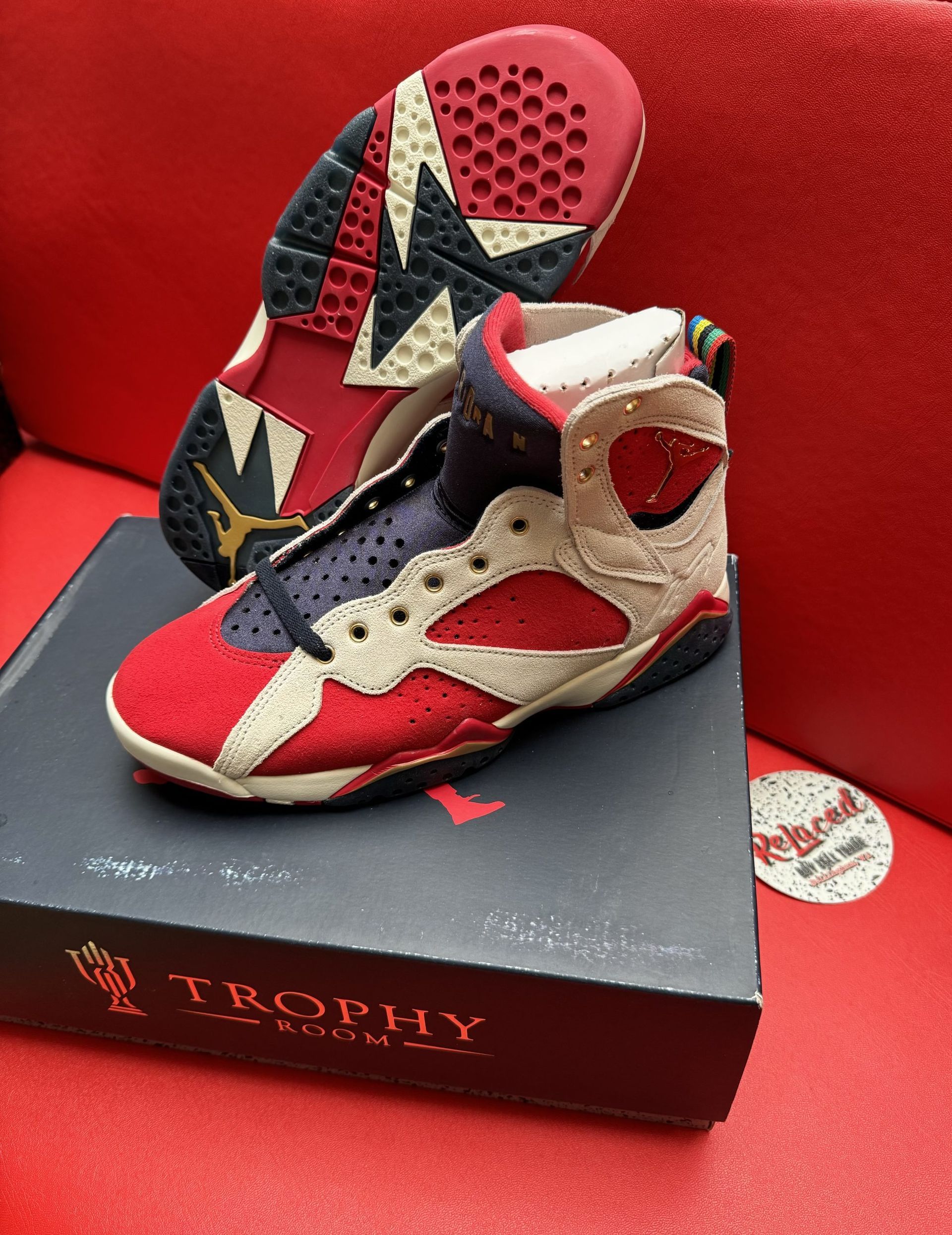 Pair of red, blue, and tan Air Jordan 7 sneakers on top of a shoe box with a red background.