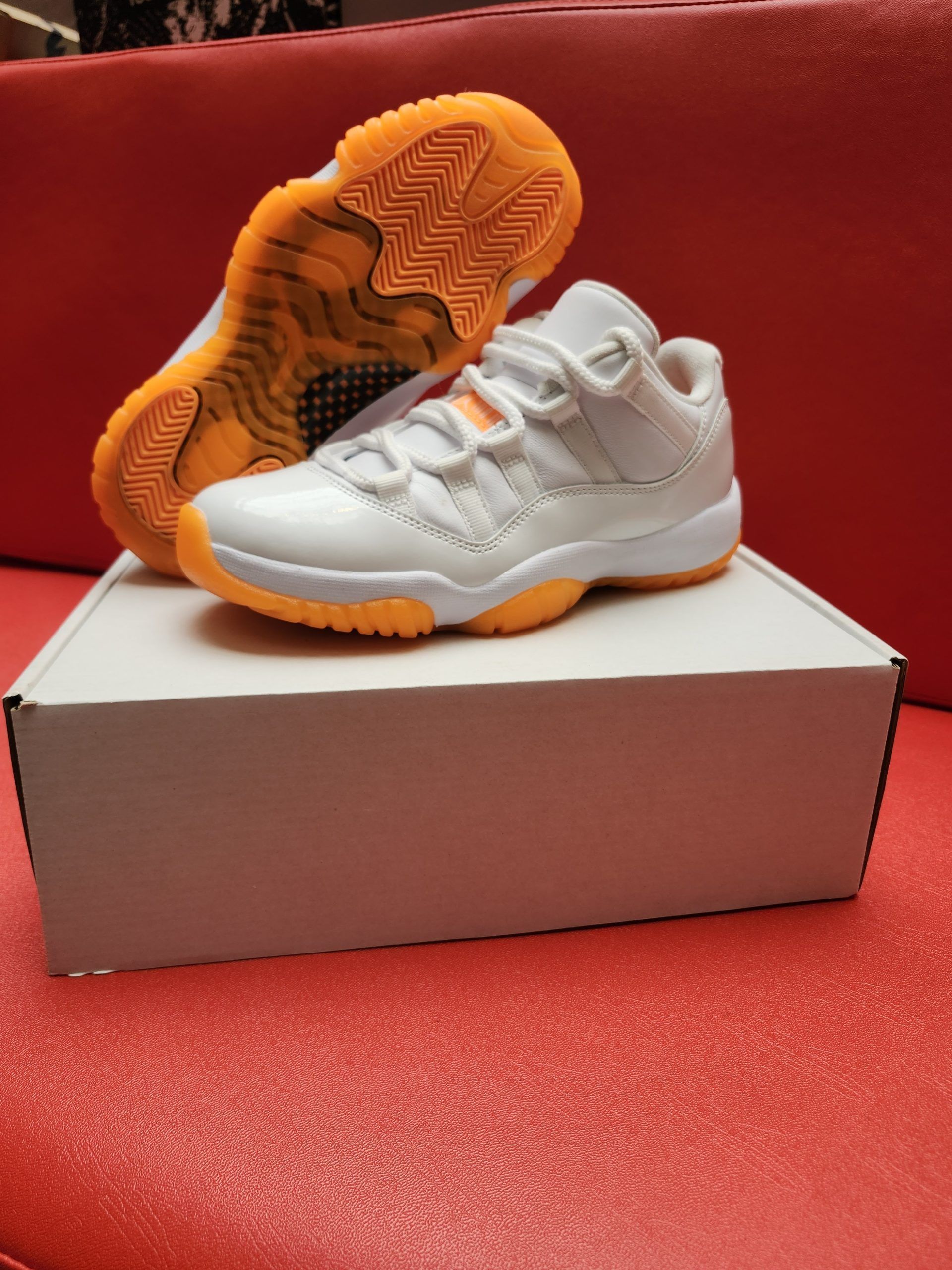 White and orange Air Jordan 11 low-top sneakers on a white box, set against a red background.