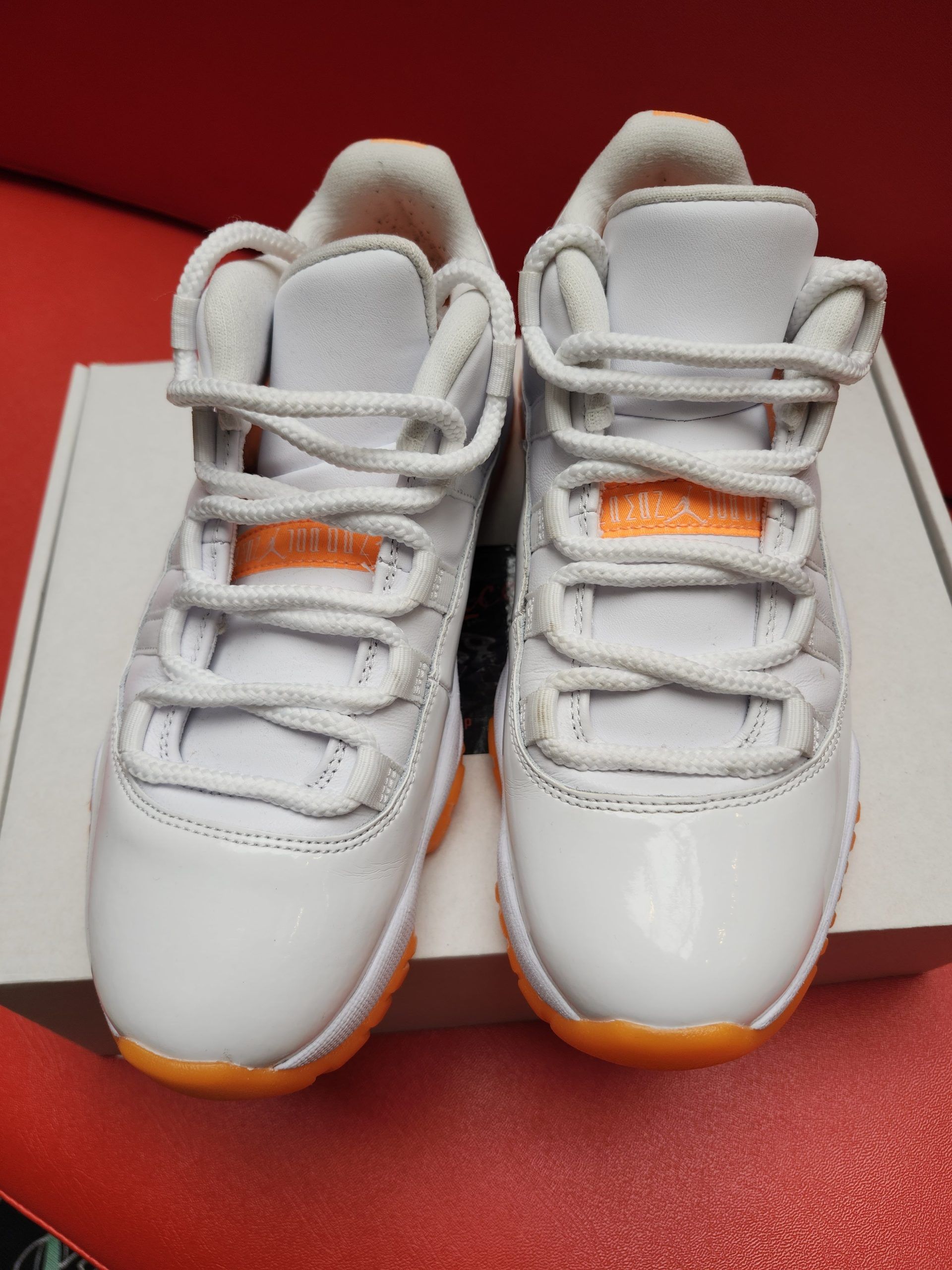 White and orange Air Jordan 11 low-top sneakers on a white box, set against a red background.