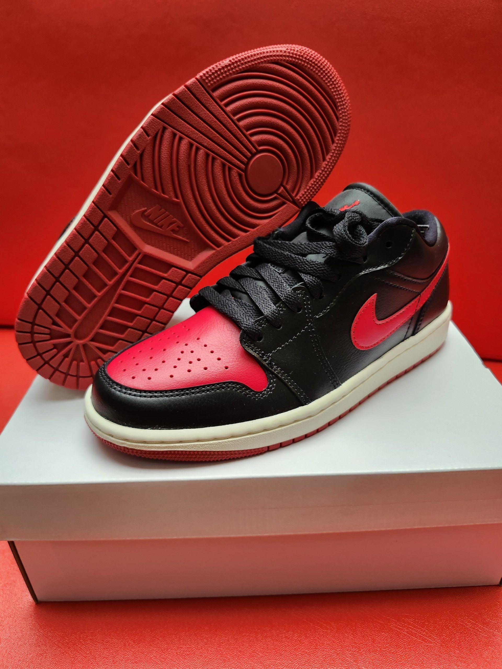 Black and red Air Jordan 1 Low sneaker with red sole and red swoosh, displayed on a white box.