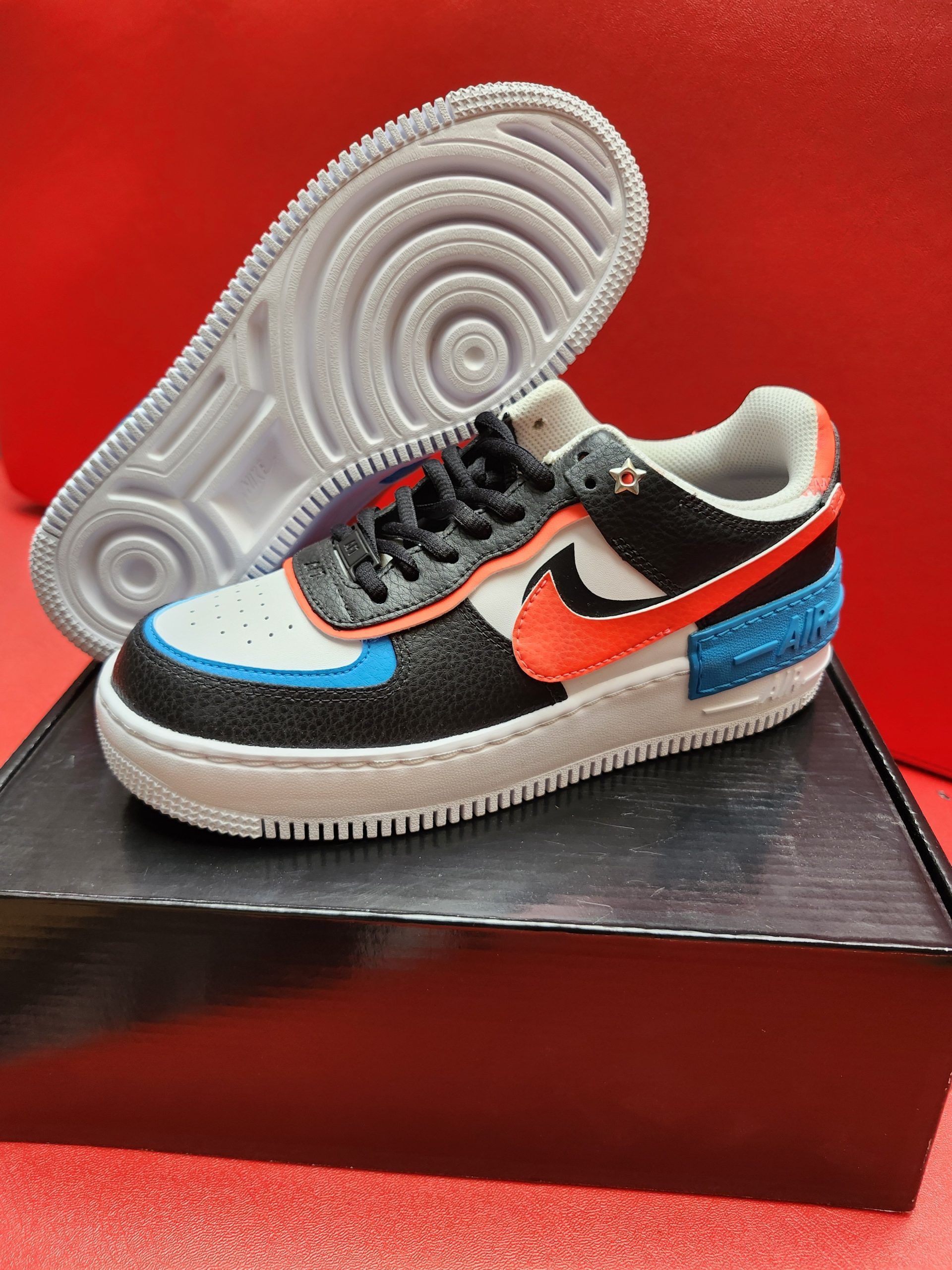 Black, orange, and white Nike Air Force 1 shoe on a black box, with the sole visible beside it on a red background.