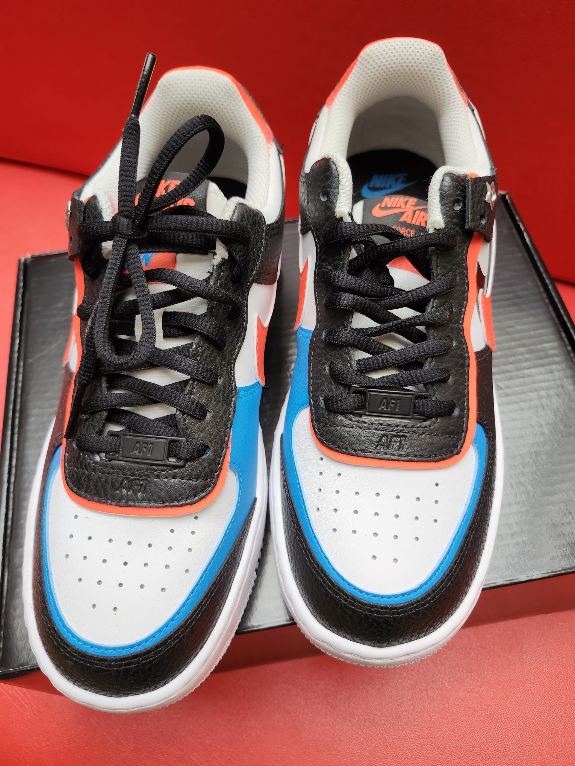 Black, orange, and white Nike Air Force 1 shoe on a black box, with the sole visible beside it on a red background.