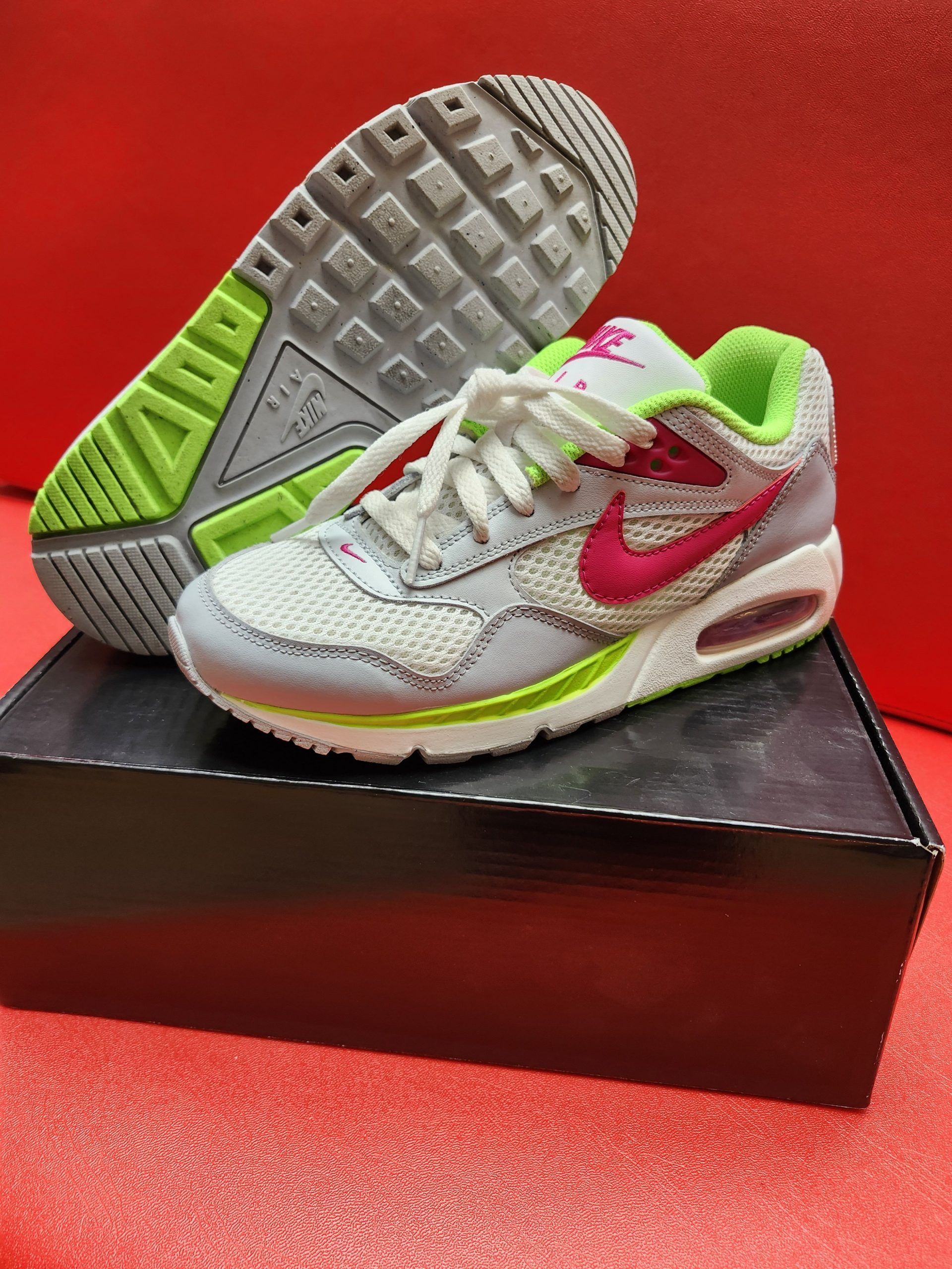 Gray and pink Nike Air Max sneakers with neon green accents on a black box. The sole of one sneaker is visible.