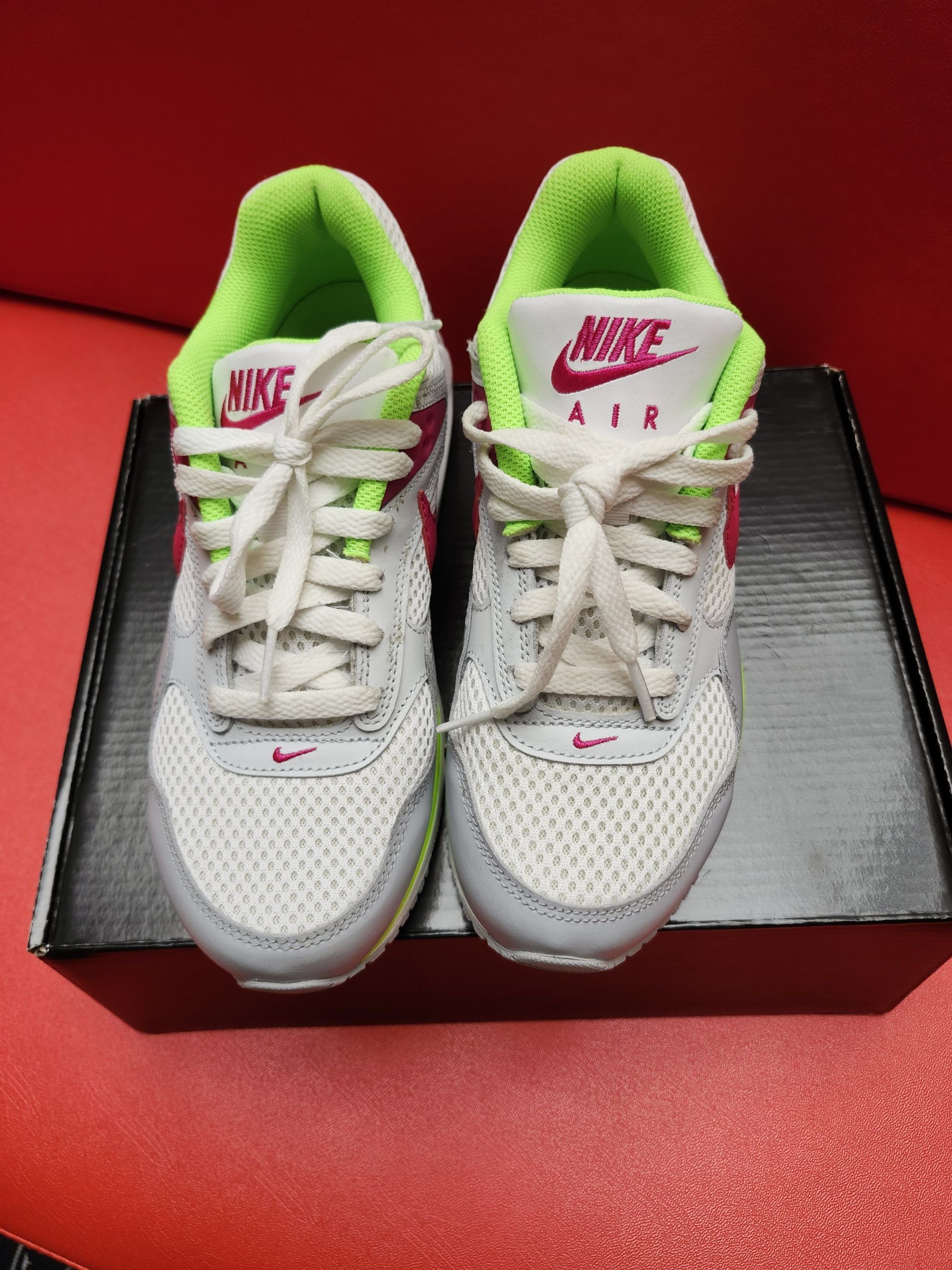Gray and pink Nike Air Max sneakers with neon green accents on a black box. The sole of one sneaker is visible.