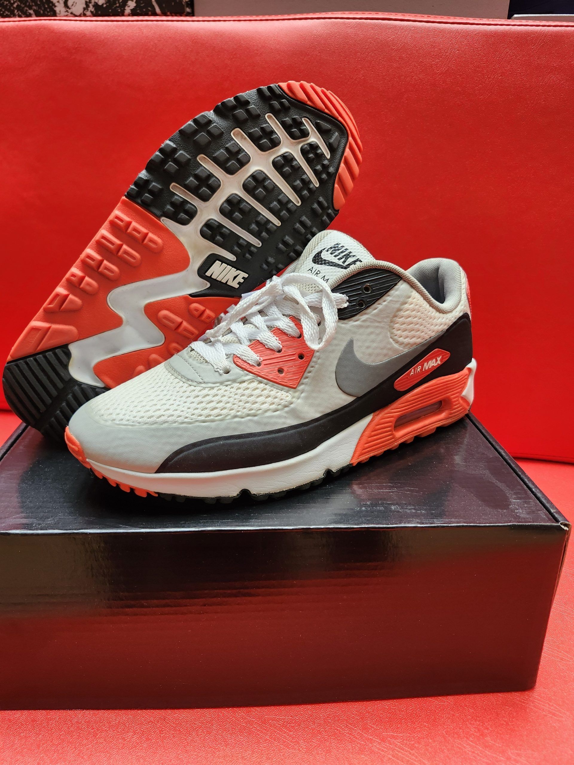 Nike Air Max 90 sneakers, white, gray, black, and orange. One shoe upright, other sole facing up, set on a black box.