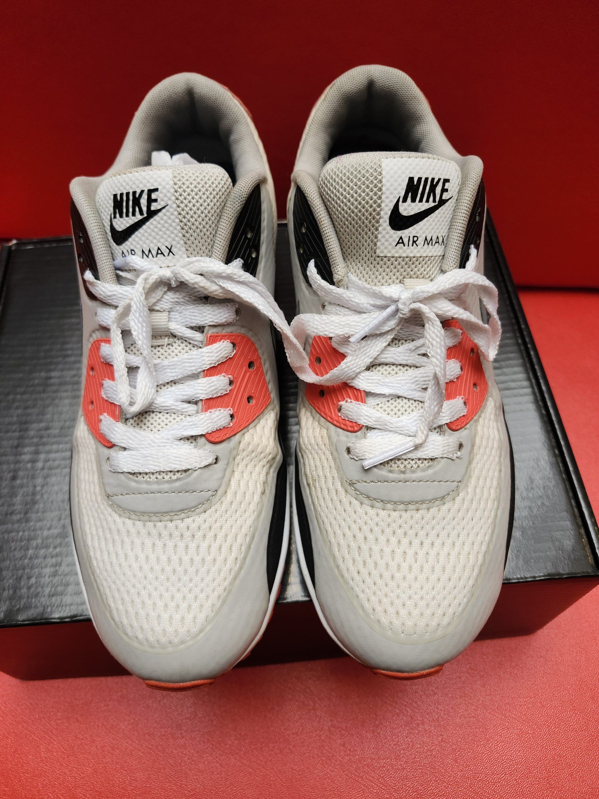 Nike Air Max 90 sneakers, white, gray, black, and orange. One shoe upright, other sole facing up, set on a black box.