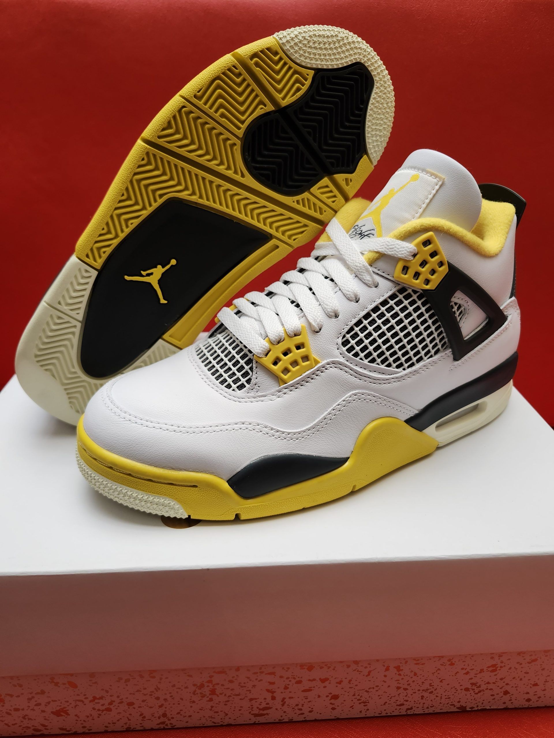 White, yellow, and black Air Jordan 4 sneaker with the sole up in the background, resting on a white surface with a red backdrop.