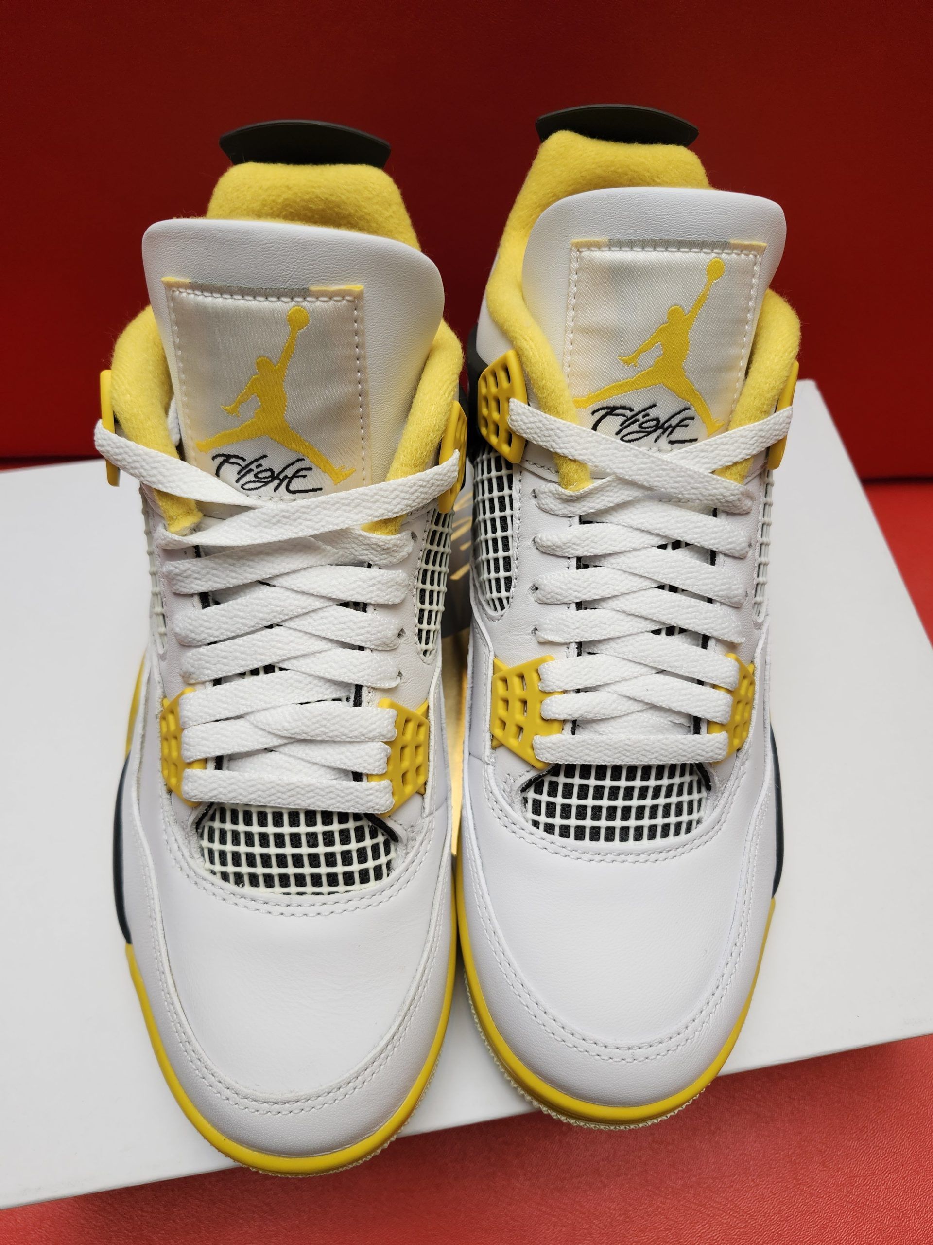 White, yellow, and black Air Jordan 4 sneaker with the sole up in the background, resting on a white surface with a red backdrop.