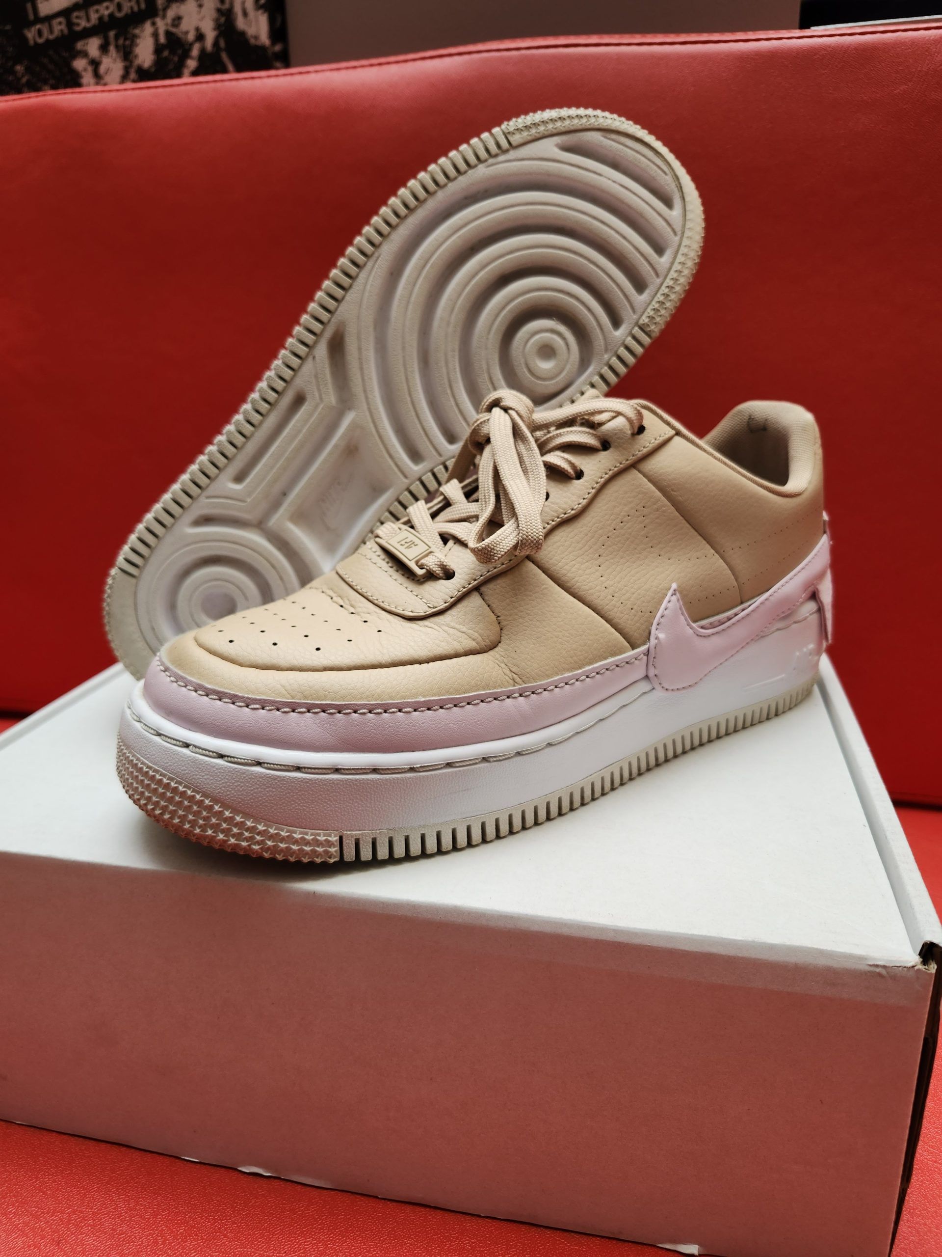 Beige and pink Nike Air Force 1 sneakers on white box, red background.