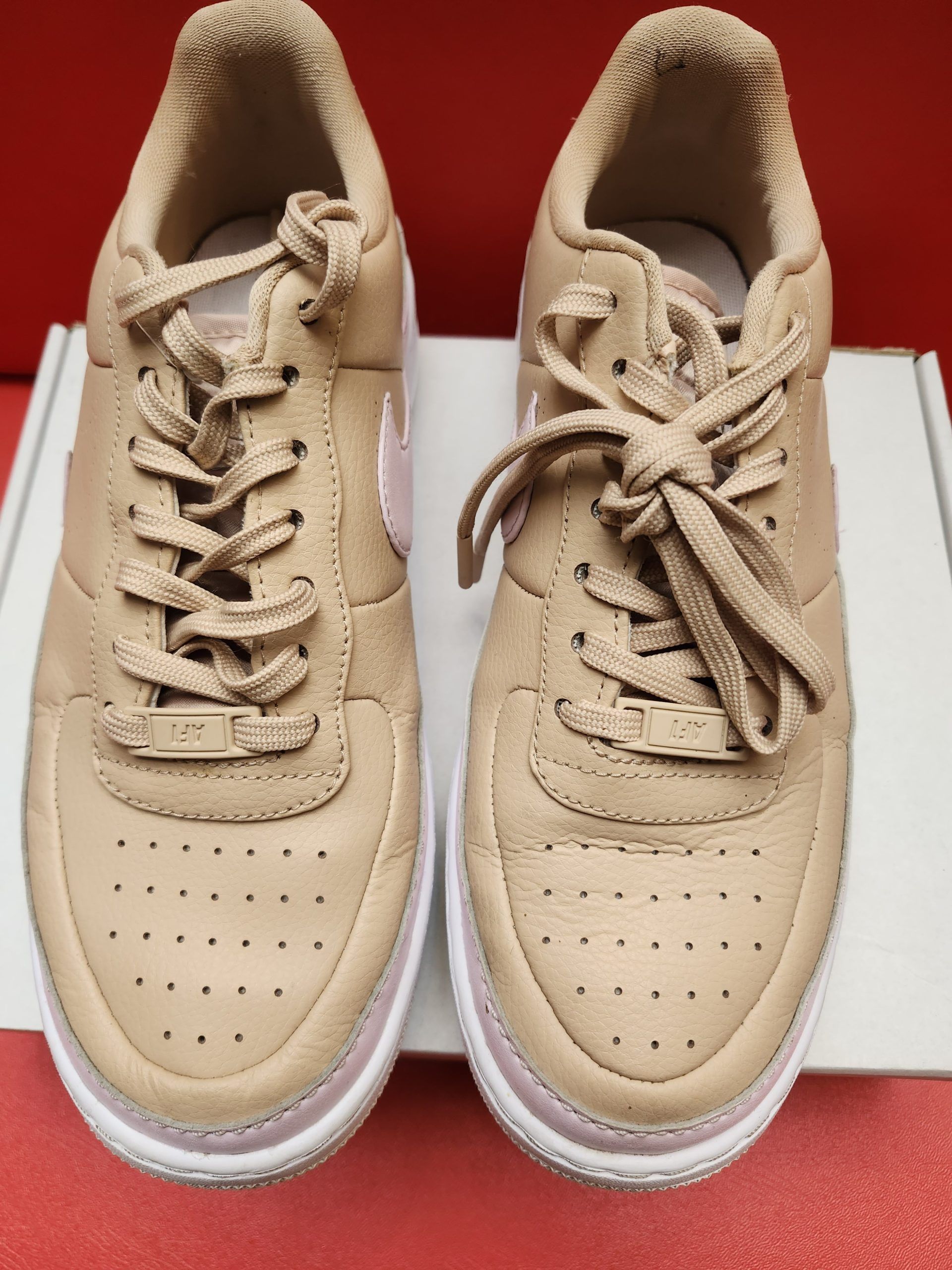 Beige and pink Nike Air Force 1 sneakers on white box, red background.