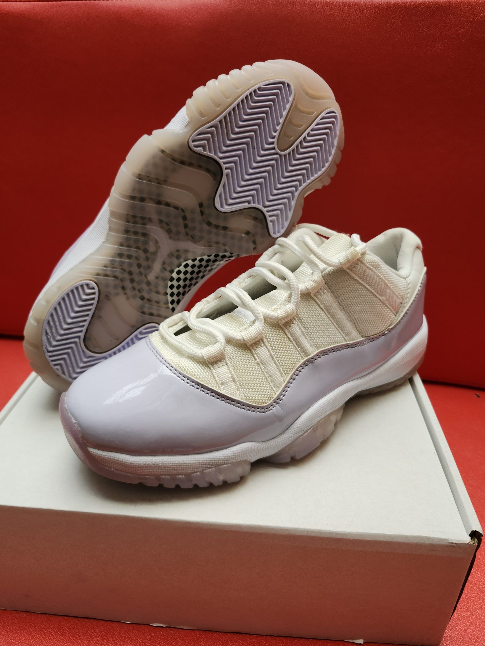 Air Jordan 11 Low sneakers, white with pale purple patent leather, resting on a white box against a red backdrop.