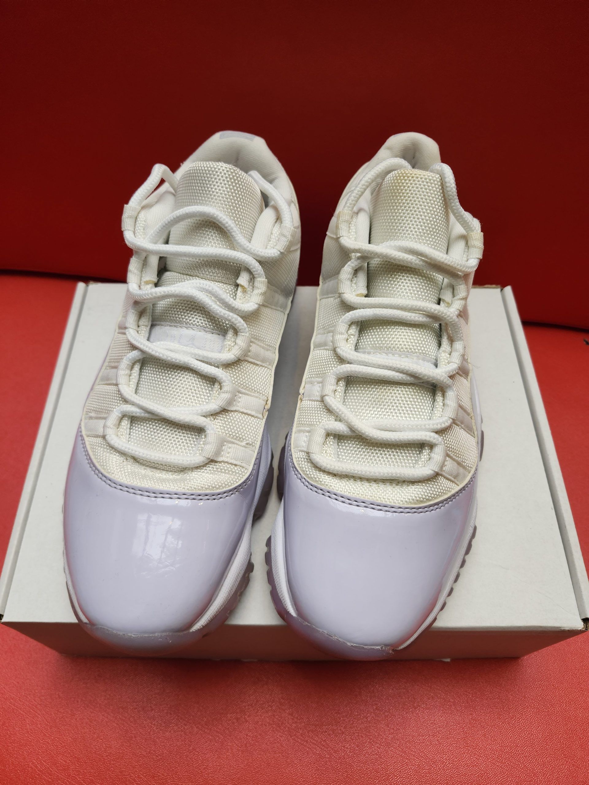 Air Jordan 11 Low sneakers, white with pale purple patent leather, resting on a white box against a red backdrop.