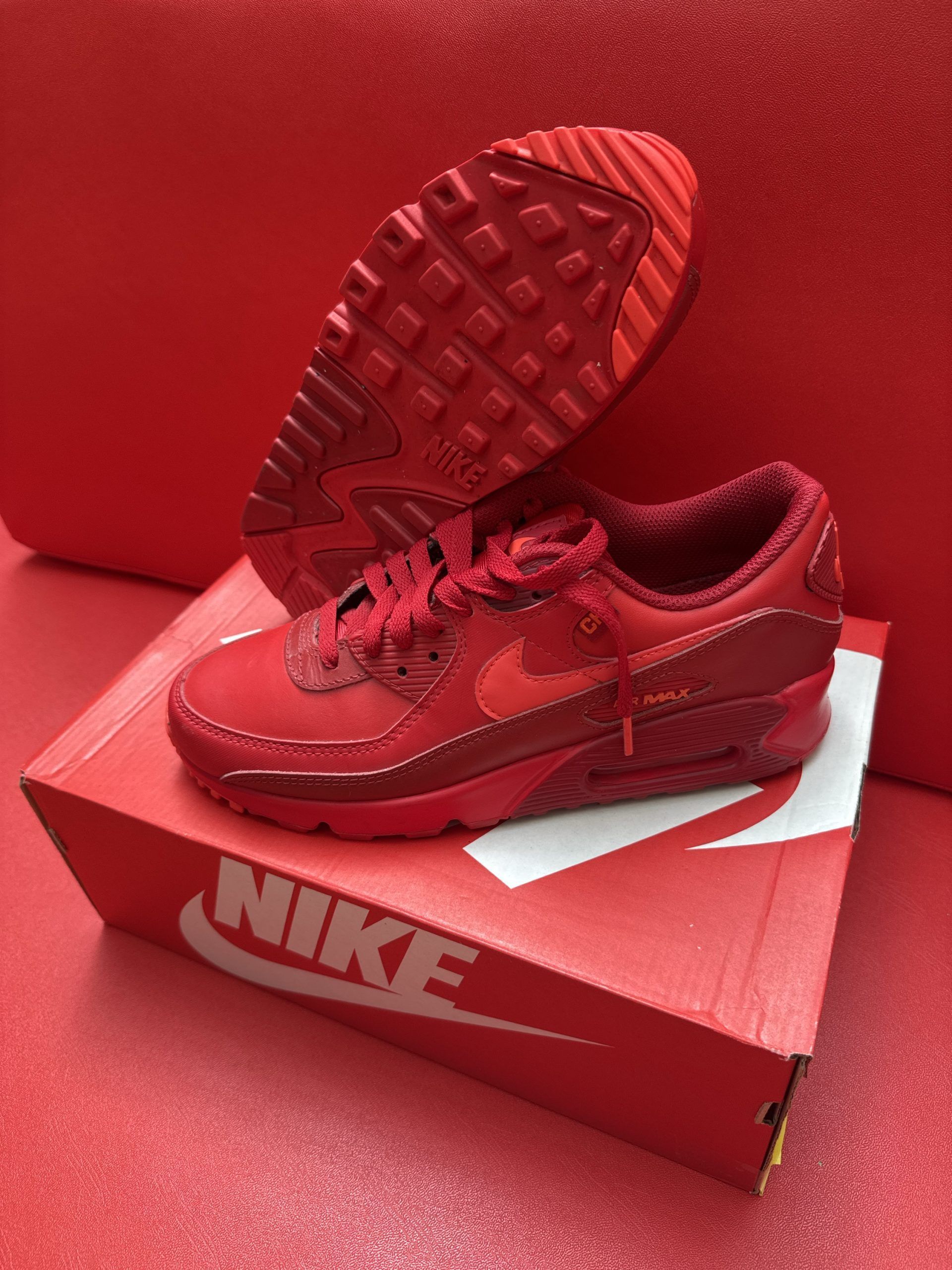 Red Nike Air Max 90 sneakers on a red Nike shoe box. One shoe upside down, showing sole.