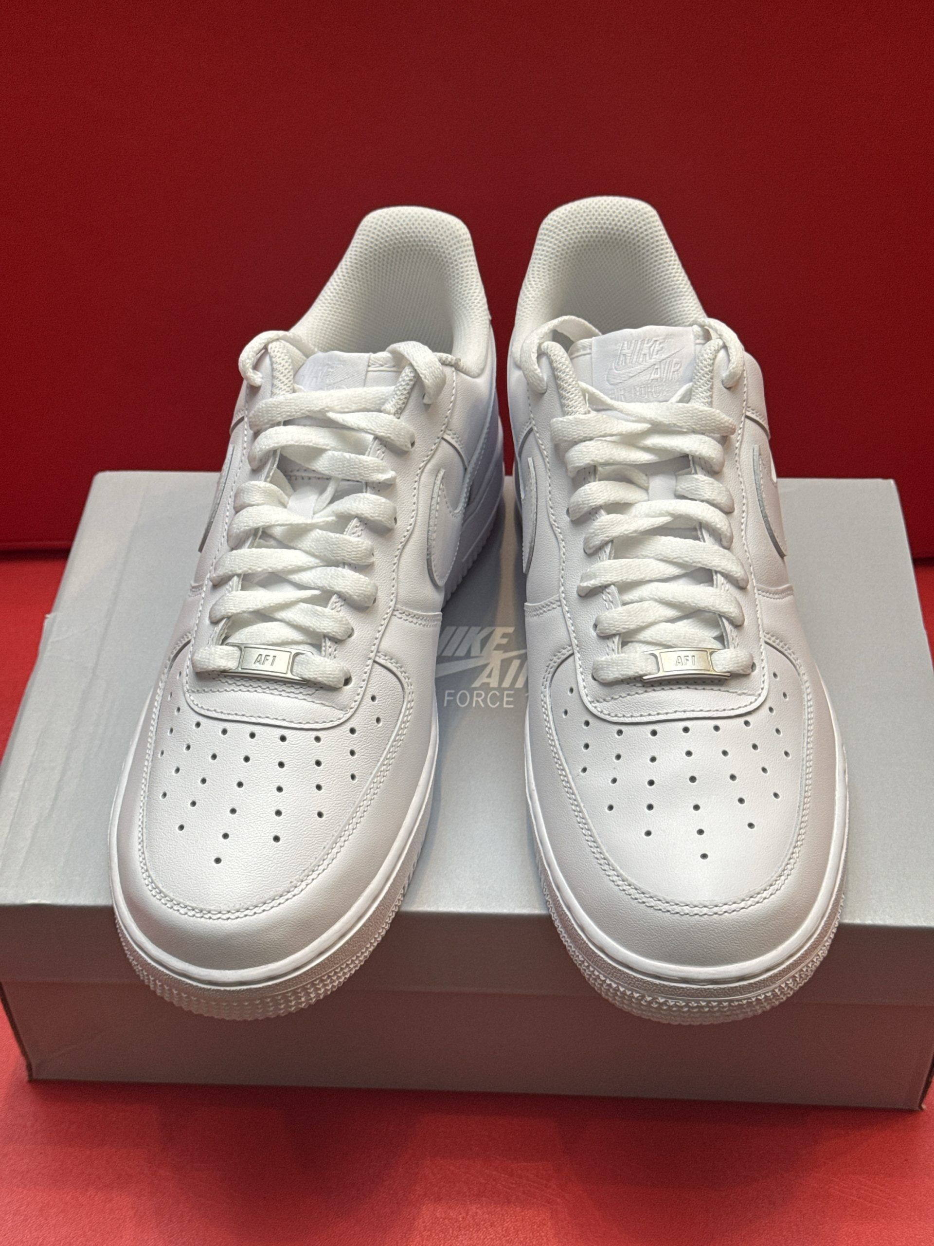 White Nike Air Force 1 sneakers on a white box, one shoe upside down showing the sole; red background.