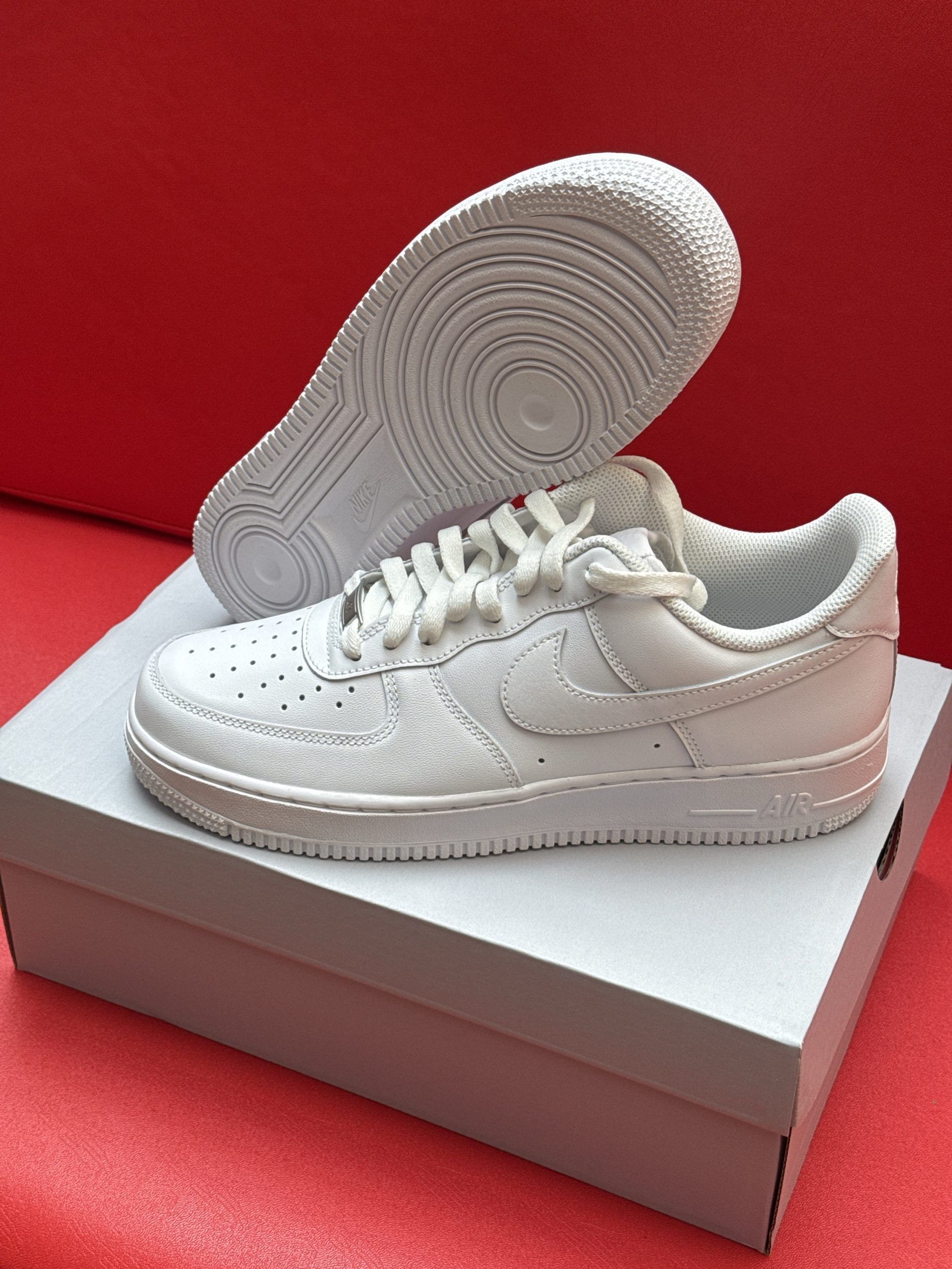 White Nike Air Force 1 sneakers on a box, sole up, against a red background.