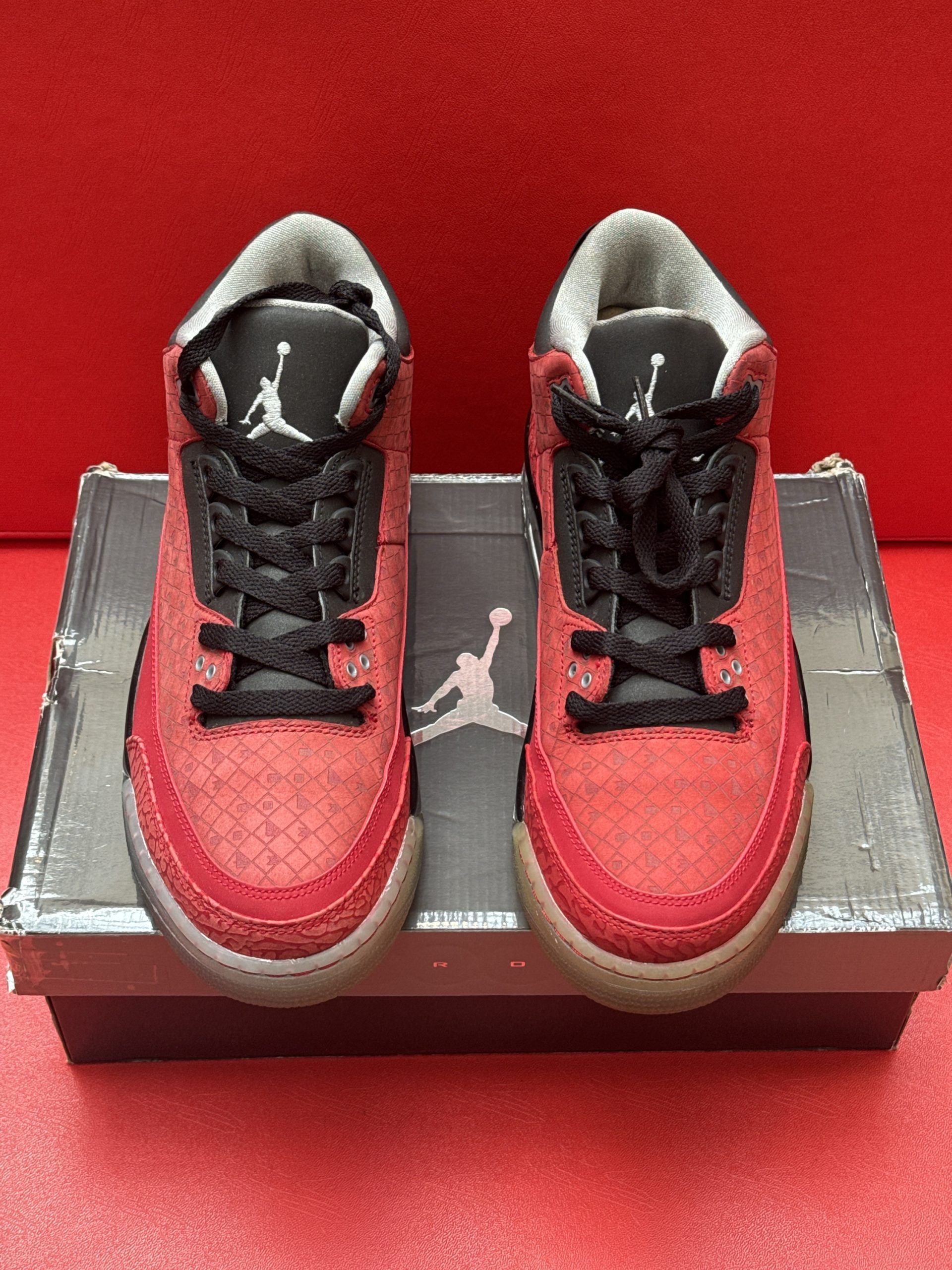 Red and black Air Jordan 3 sneakers with a worn sole on a shoebox against a red background.