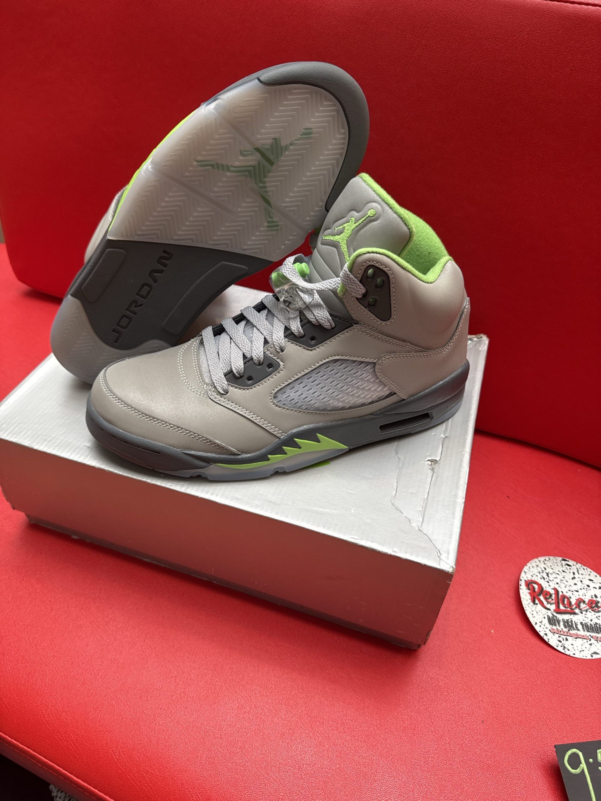 Gray and green Air Jordan 5 sneakers on a white box, sole visible, set on a red surface.