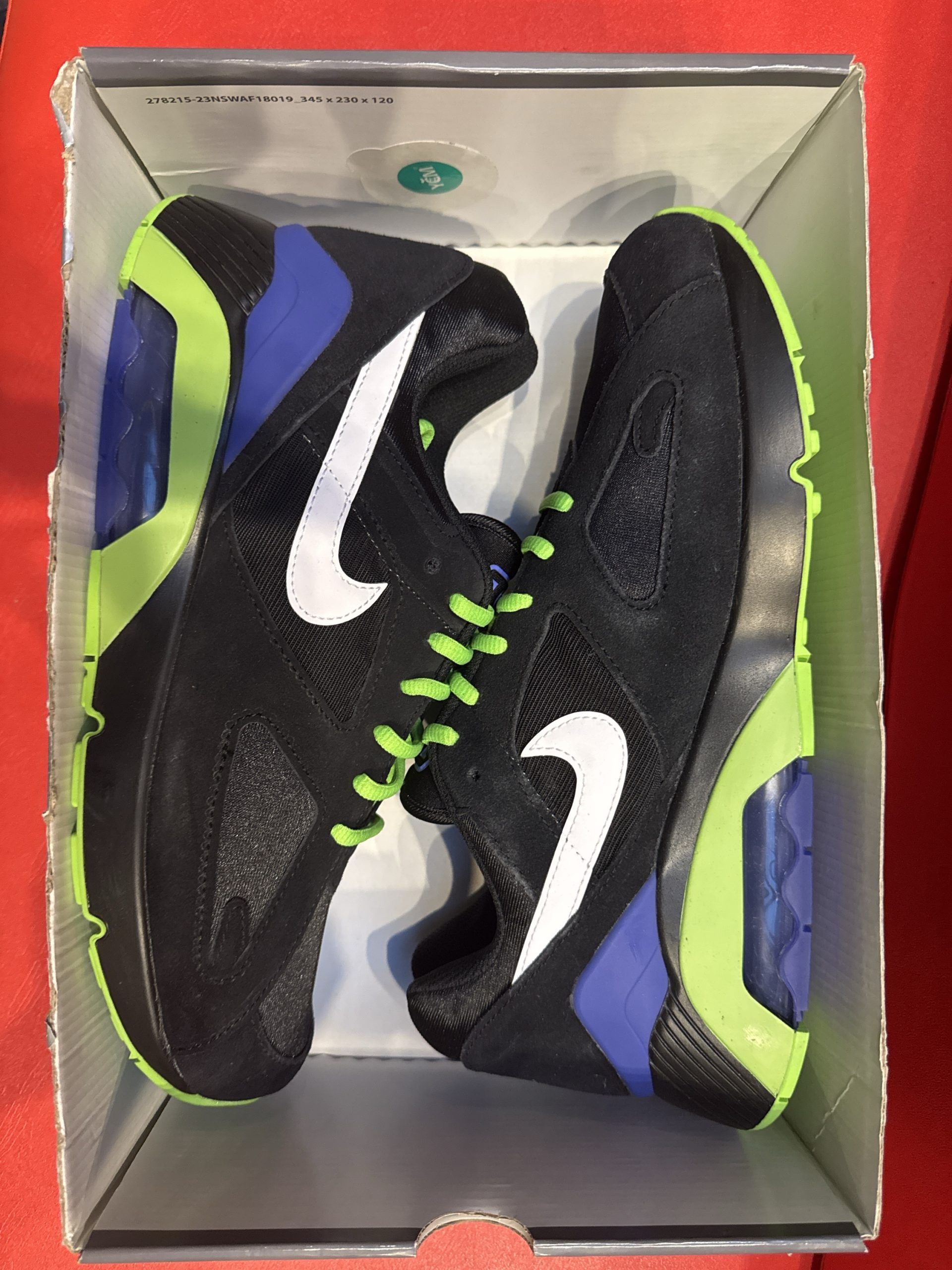 Black Nike Air 180 sneakers with a white swoosh, sitting on a shoebox. Green and blue accents.