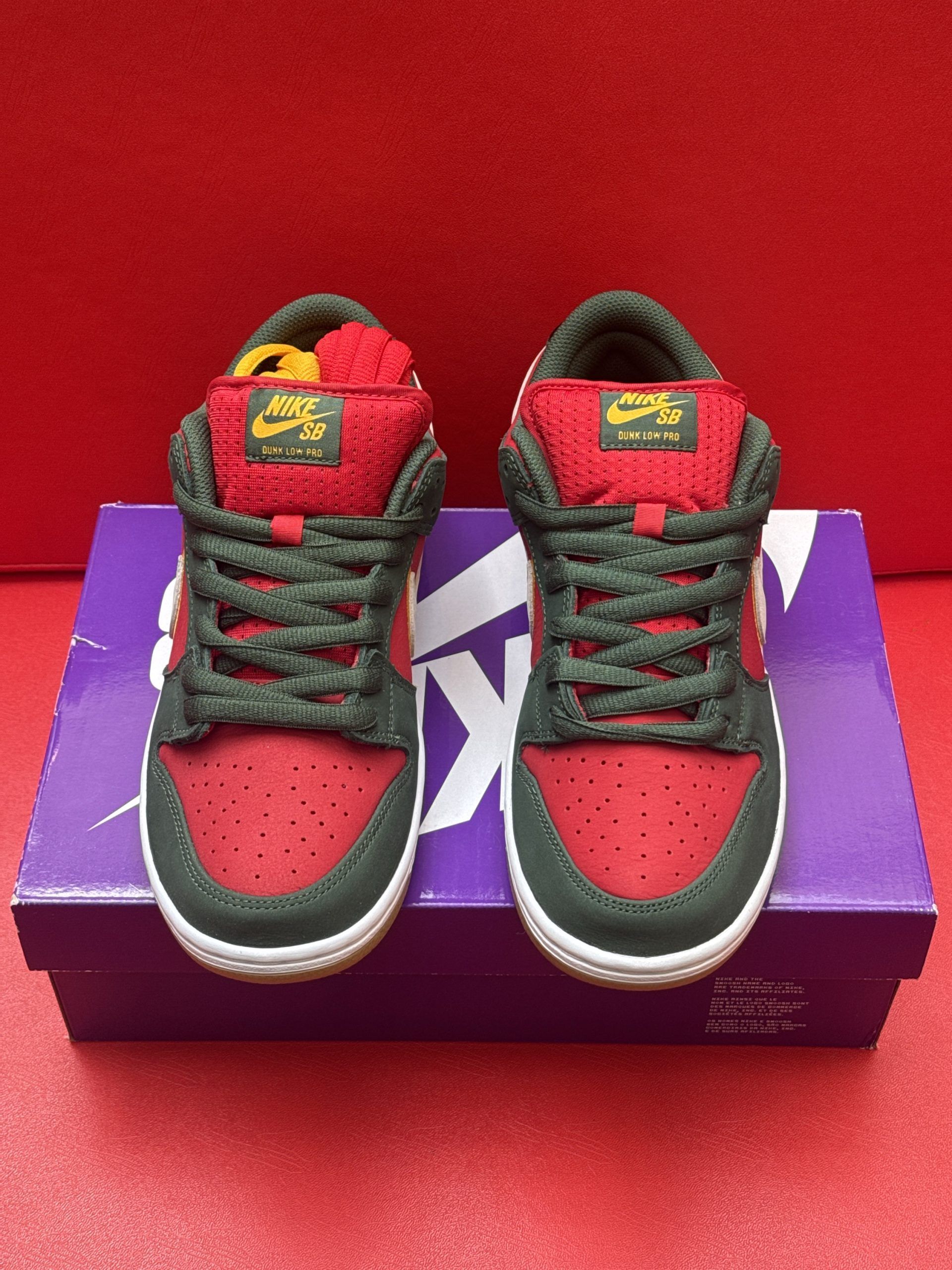 Nike SB Dunk Low sneakers, red, black, and white, on top of a purple shoebox with a brown sole on a red surface.