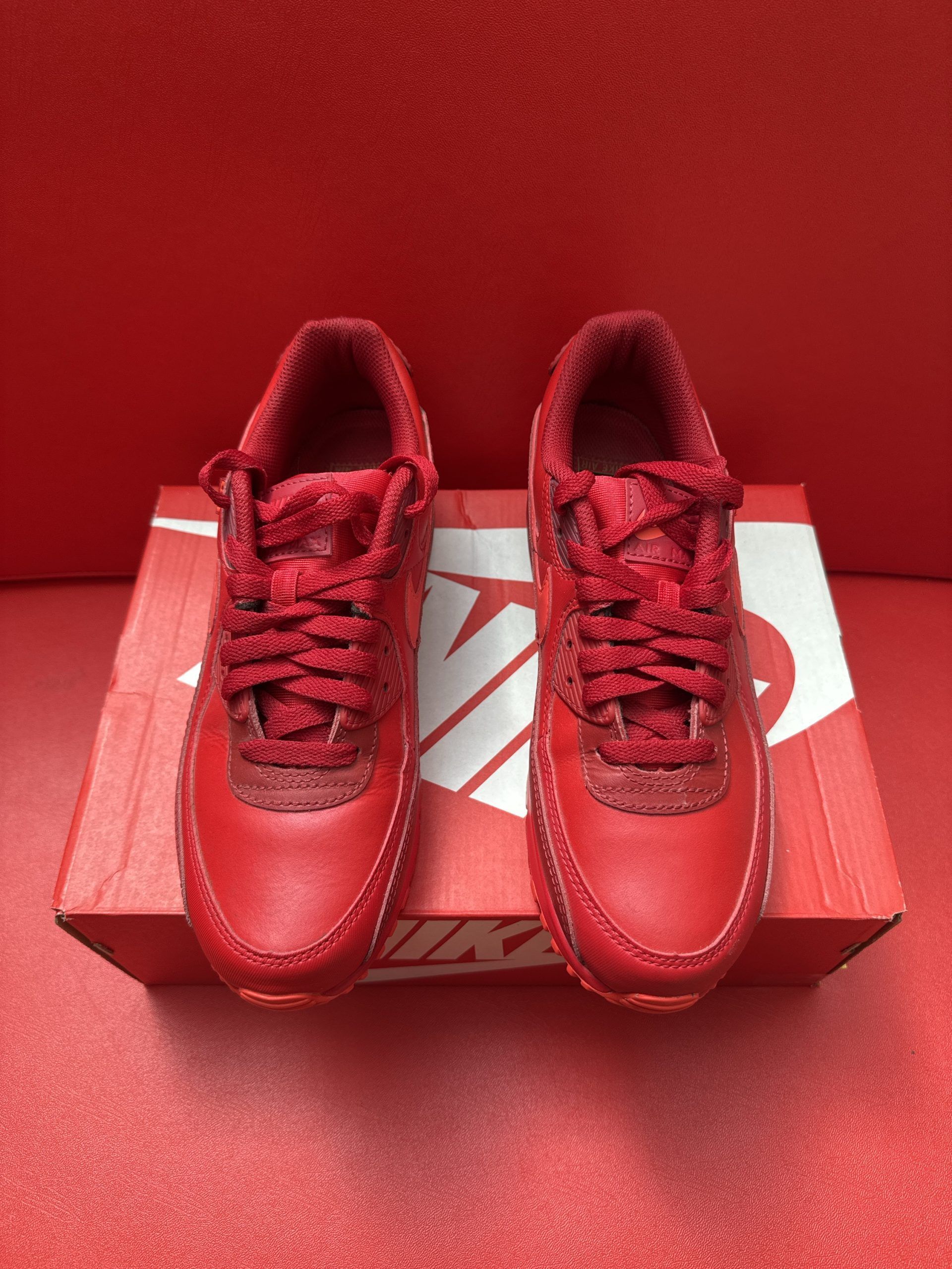 Red Nike Air Max 90 sneakers on a red Nike shoe box. One shoe upside down, showing sole.